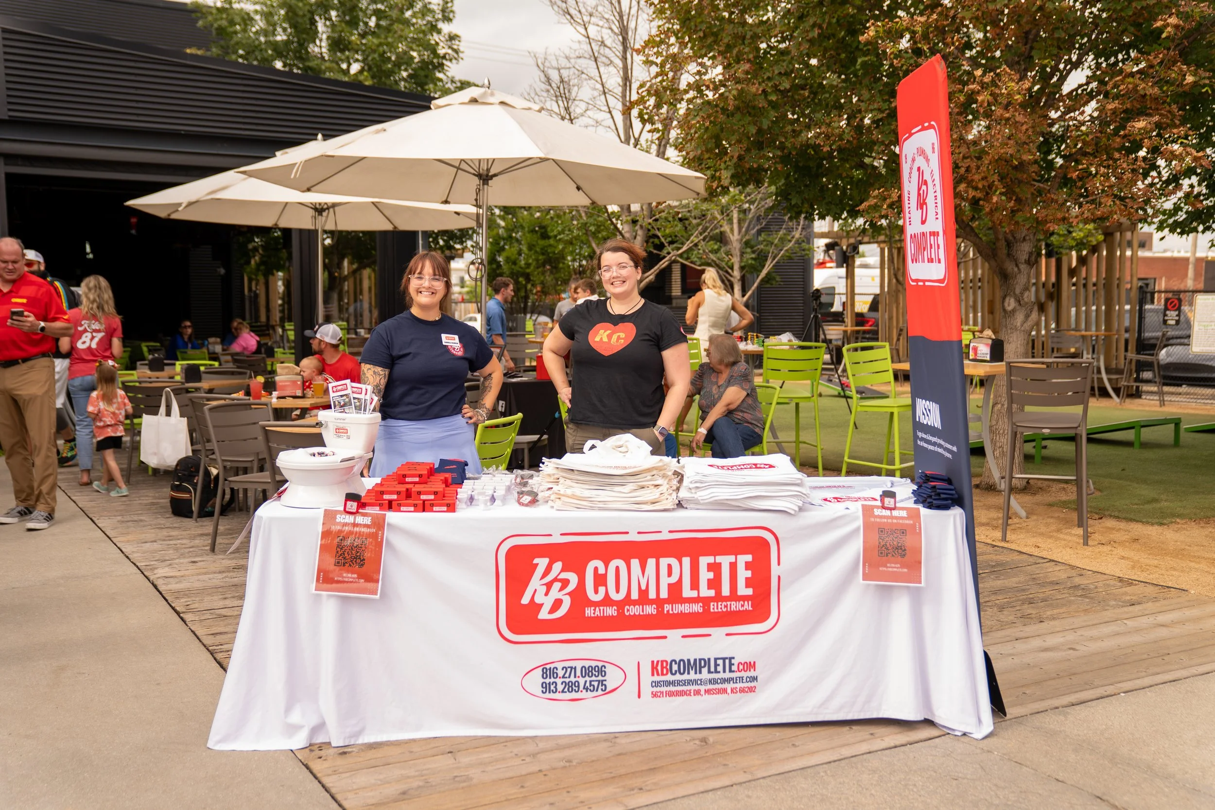 Two women standing behind a table at an outdoor event, promoting KB Complete, a heating, cooling, plumbing, and electrical company. The table is covered with promotional materials, and there is a QR code on a sign for scanning. Other people are visib