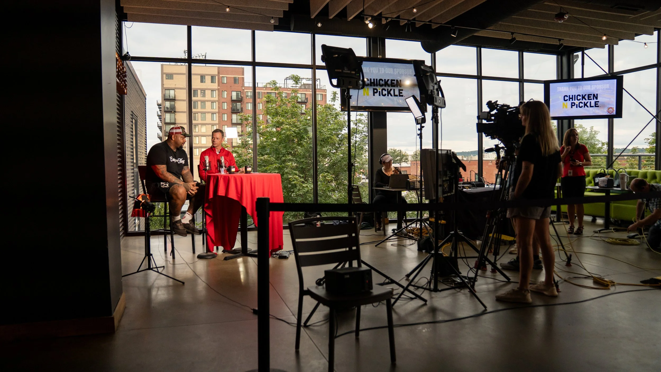 Recording studio with two men sitting at a table with microphones, large windows showing buildings outside, and camera crew filming them, with two screens displaying "CHICKEN N PICKLE".