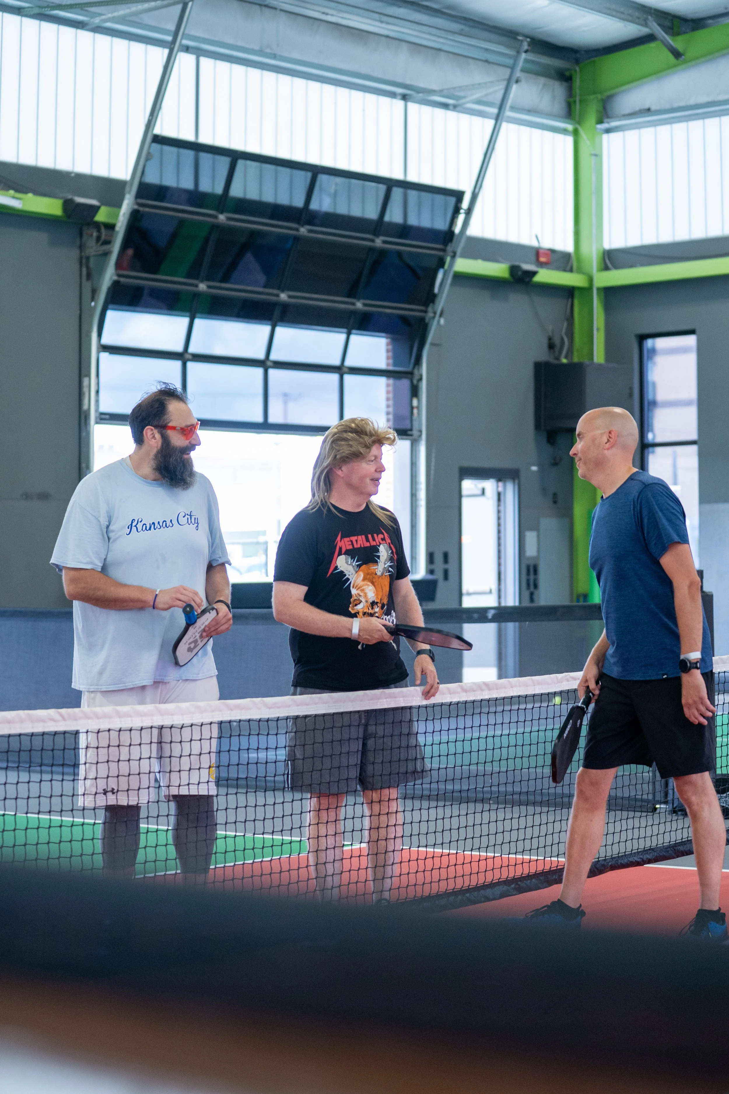 Three men standing on a pickleball court inside a gym, chatting after a game. One is holding a paddle, another wears a Kansas City t-shirt, and the third, in a Metallica t-shirt, is holding a paddle and a ball.