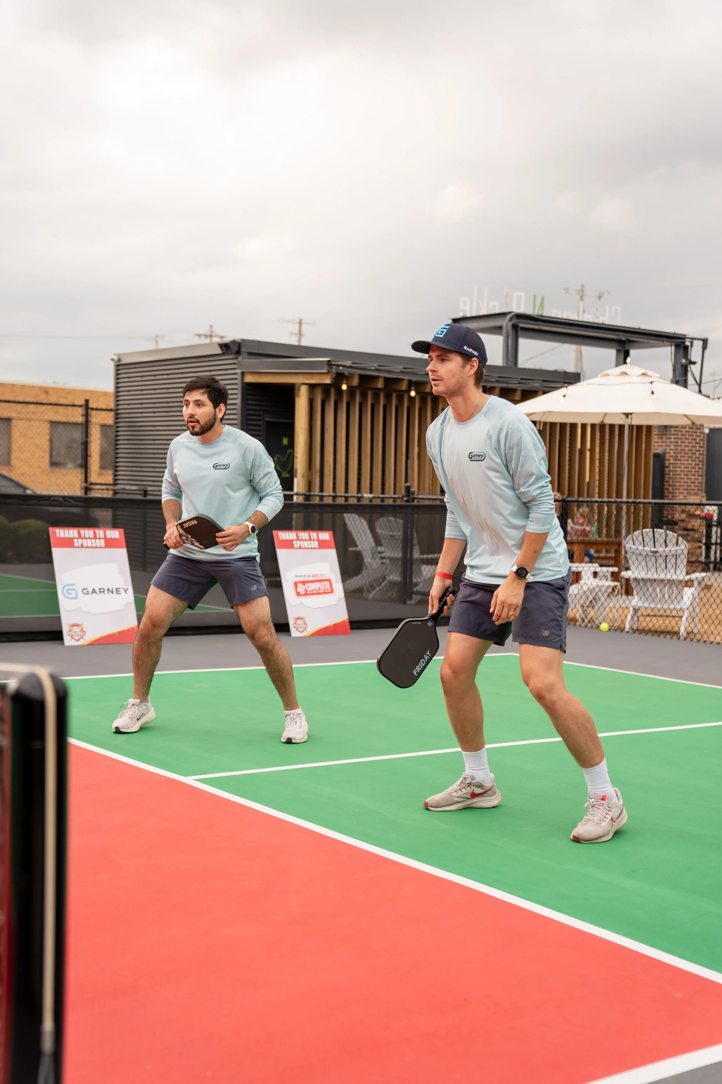 Two men playing pickleball on an outdoor court with a black fence, signs, and outdoor furniture in background, overcast sky.