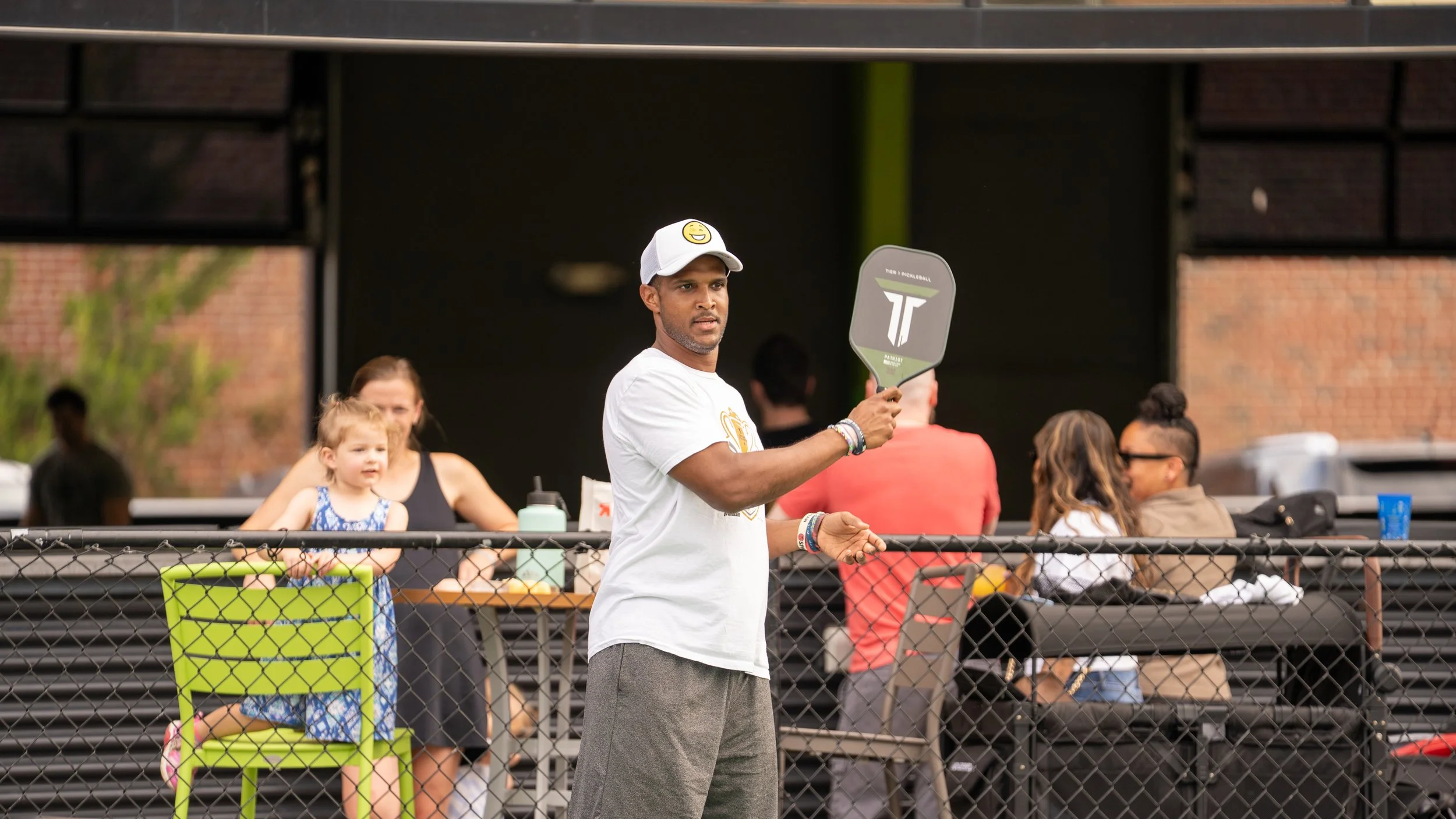 A man holding a tennis paddle stands behind a chain-link fence with a family sitting on benches in the background.