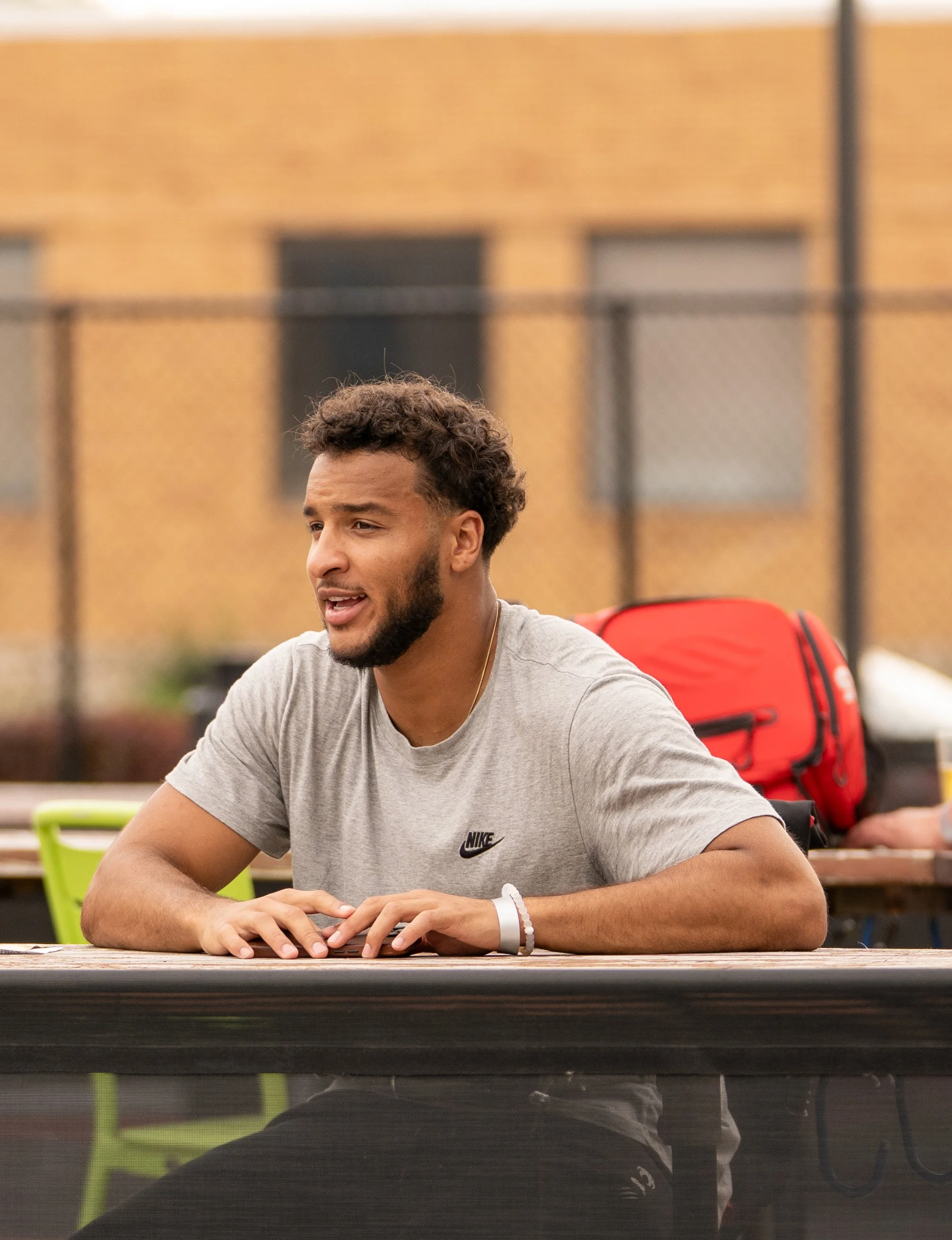 A young man with curly hair and a beard, wearing a gray Nike T-shirt, sitting at an outdoor table, talking or explaining, with a red backpack in the background.