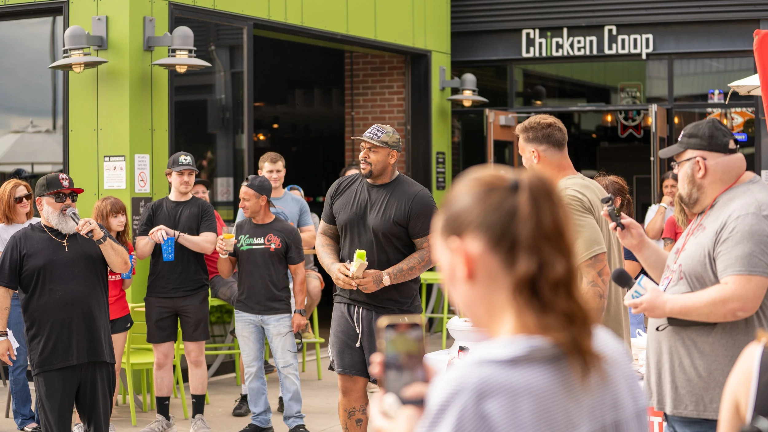 Group of people gathered outside a restaurant called 'Chicken Coop,' with a man speaking into a microphone, holding a drink and wearing sunglasses, surrounded by casually dressed individuals, some with drinks, during daytime.