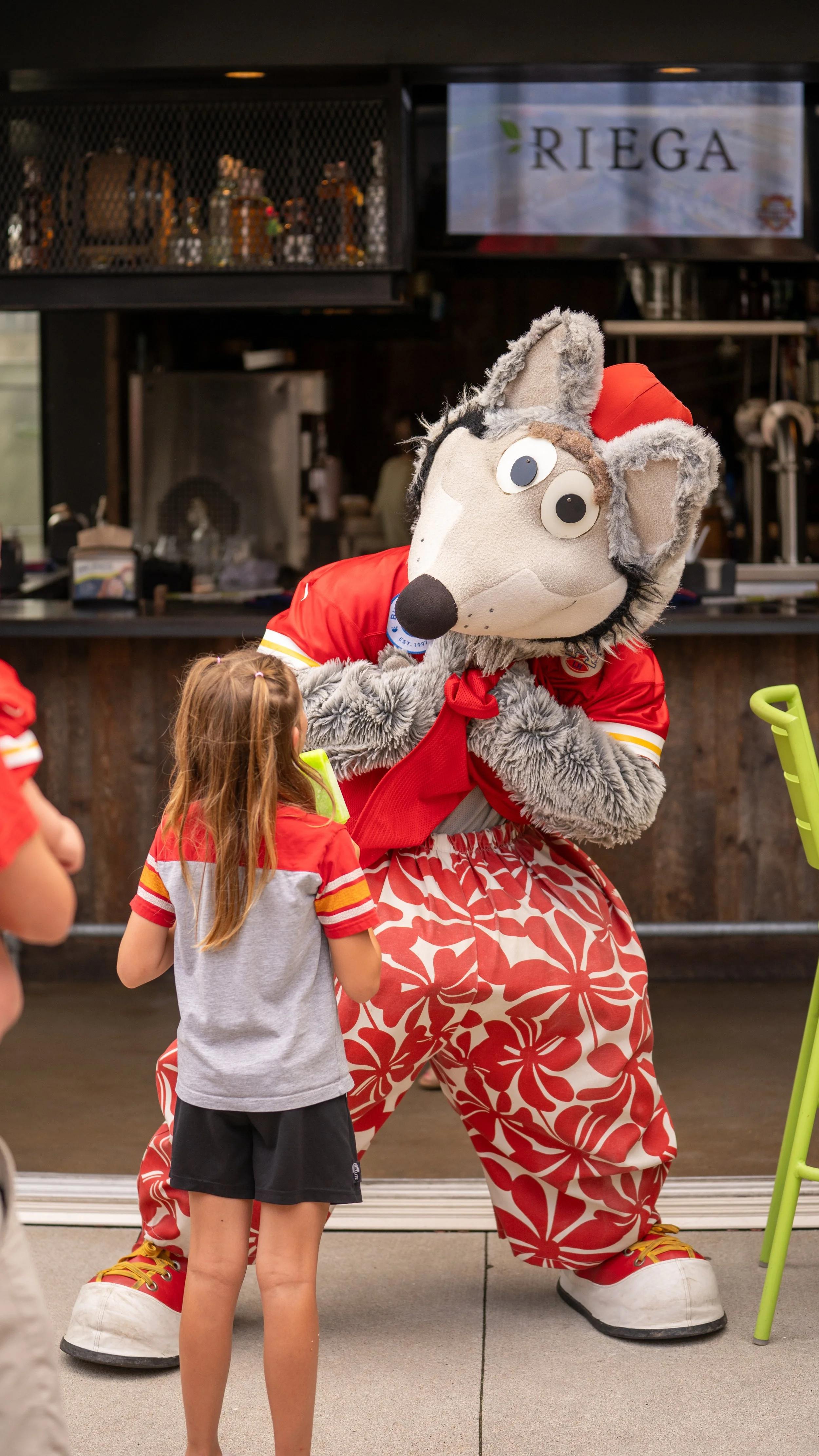 Person dressed as a mascot wolf character in red and white clothing interacting with young girl at an event.