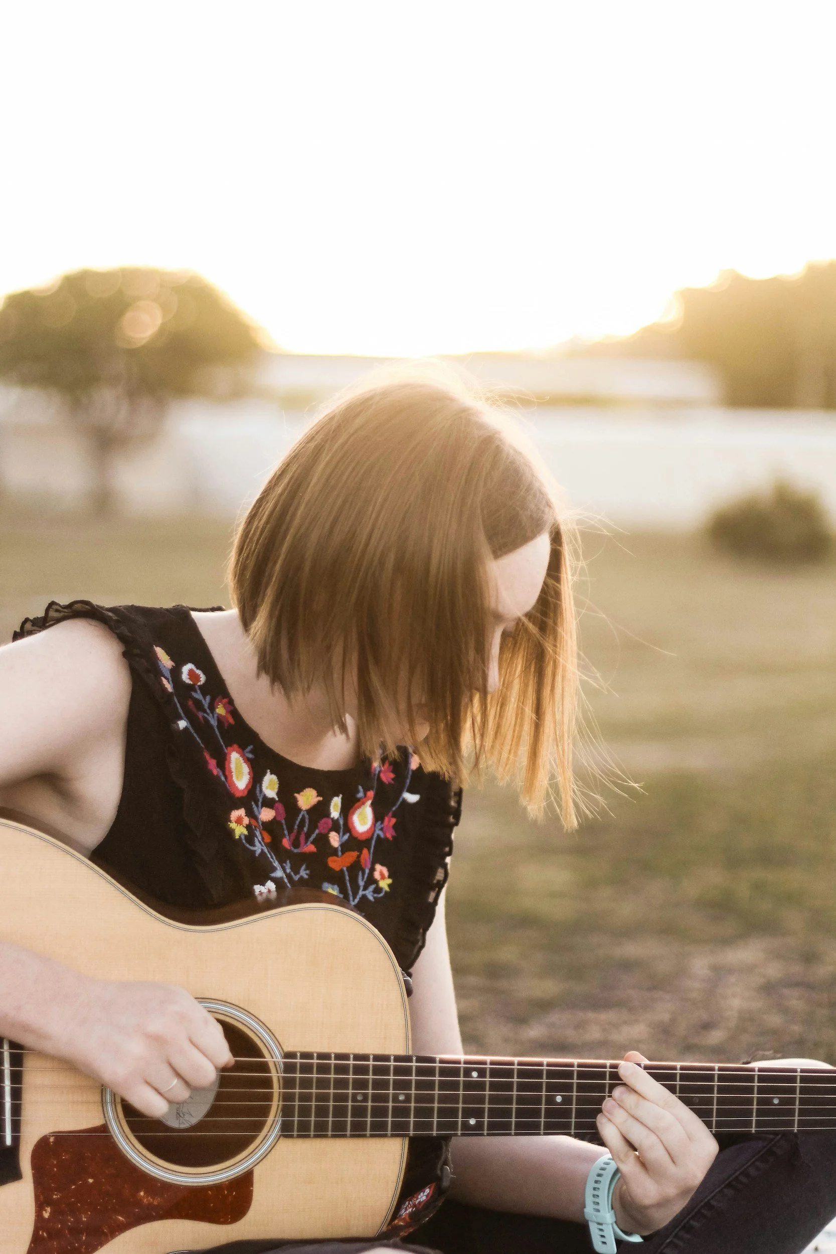 A young woman with shoulder-length red hair playing an acoustic guitar outdoors at sunset.