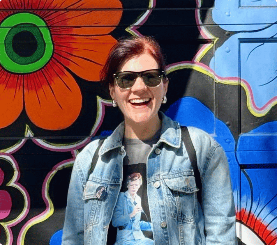 Woman with short red hair, sunglasses, and denim jacket smiling in front of a colorful, floral mural.