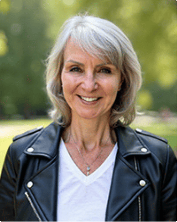 A smiling middle-aged woman with shoulder-length gray hair outdoors, wearing a black leather jacket over a white shirt, with a blurred green background.