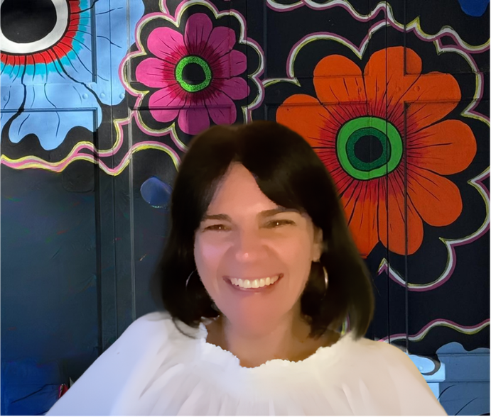 Woman with short red hair, sunglasses, and denim jacket smiling in front of a colorful, floral mural.