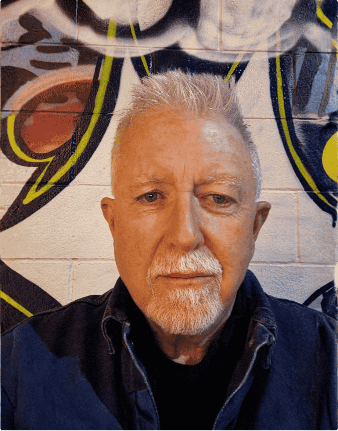 Close-up of an older man with white hair and beard, wearing a dark jacket, standing in front of a colorful mural of a cartoon character.