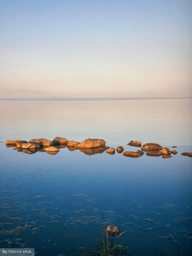 Calm lake scene with warm-toned rocks reflected in still blue water beneath a soft pastel sky.