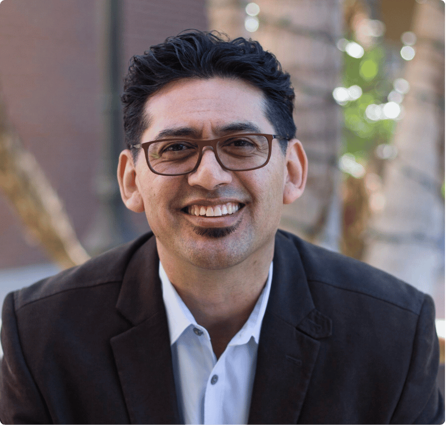 A smiling man with dark curly hair, glasses, wearing a dark blazer and white shirt, sitting outdoors with blurred trees in the background.