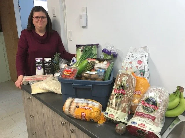 Hélène Gervais, responsable des ateliers, debout à côté d'une table avec des produits alimentaires, légumes, pains, bananes, et autres courses.