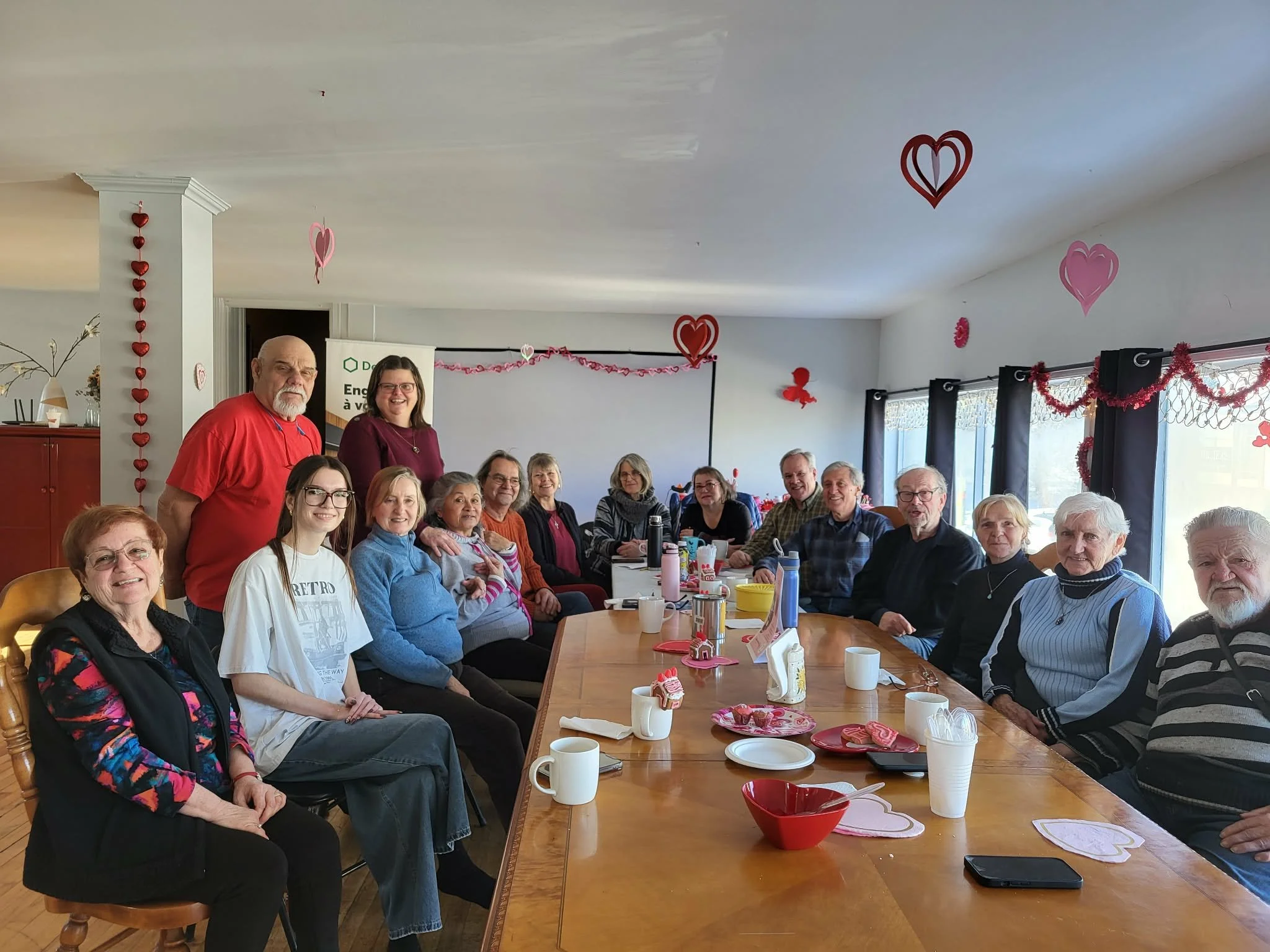 Groupe de personnes âgées et jeunes adultes réunies autour d'une grande table lors d'une fête de Saint-Valentin, décorée avec des cœurs rouges et roses suspendus au plafond, des guirlandes roses, des assiettes et des tasses fuchsia sur la table.