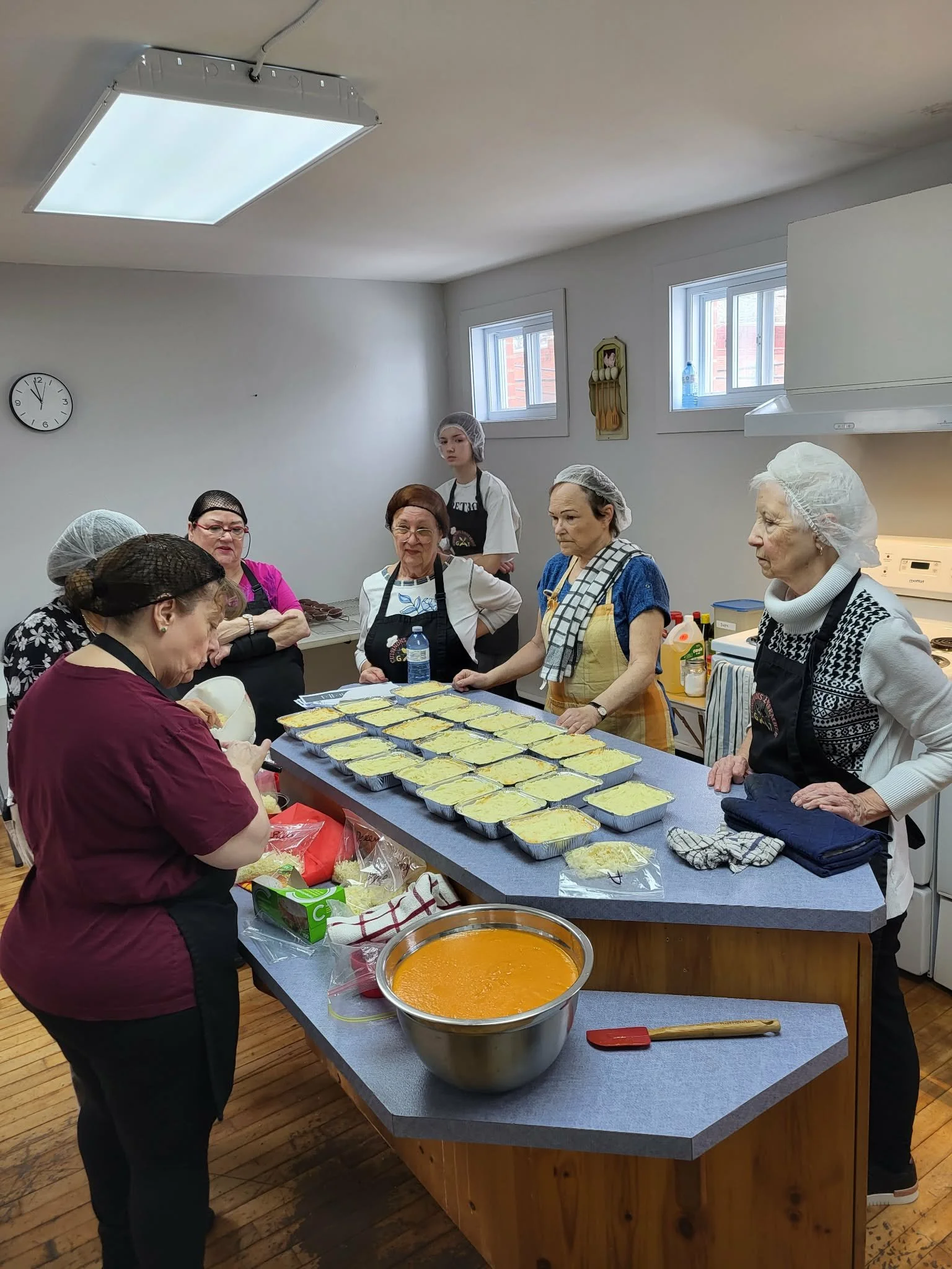 Groupe de personnes âgées dans une cuisine, portant des coiffes de cuisine, préparant et emballant des plats, avec une grande marmite sur la table.