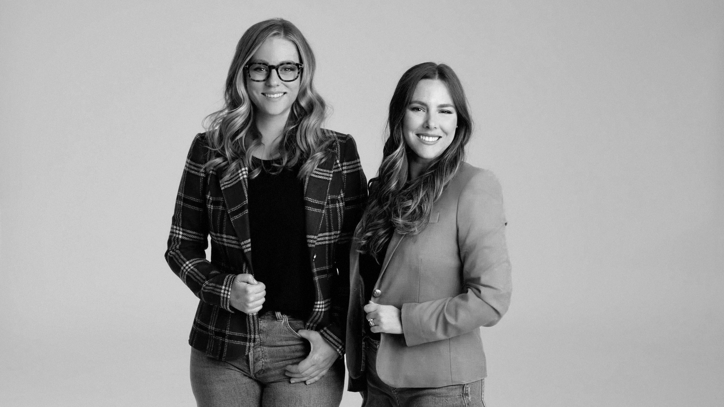 Two women smiling, standing side by side against a plain background, one wearing glasses and a plaid jacket, the other in a blazer and blouse.