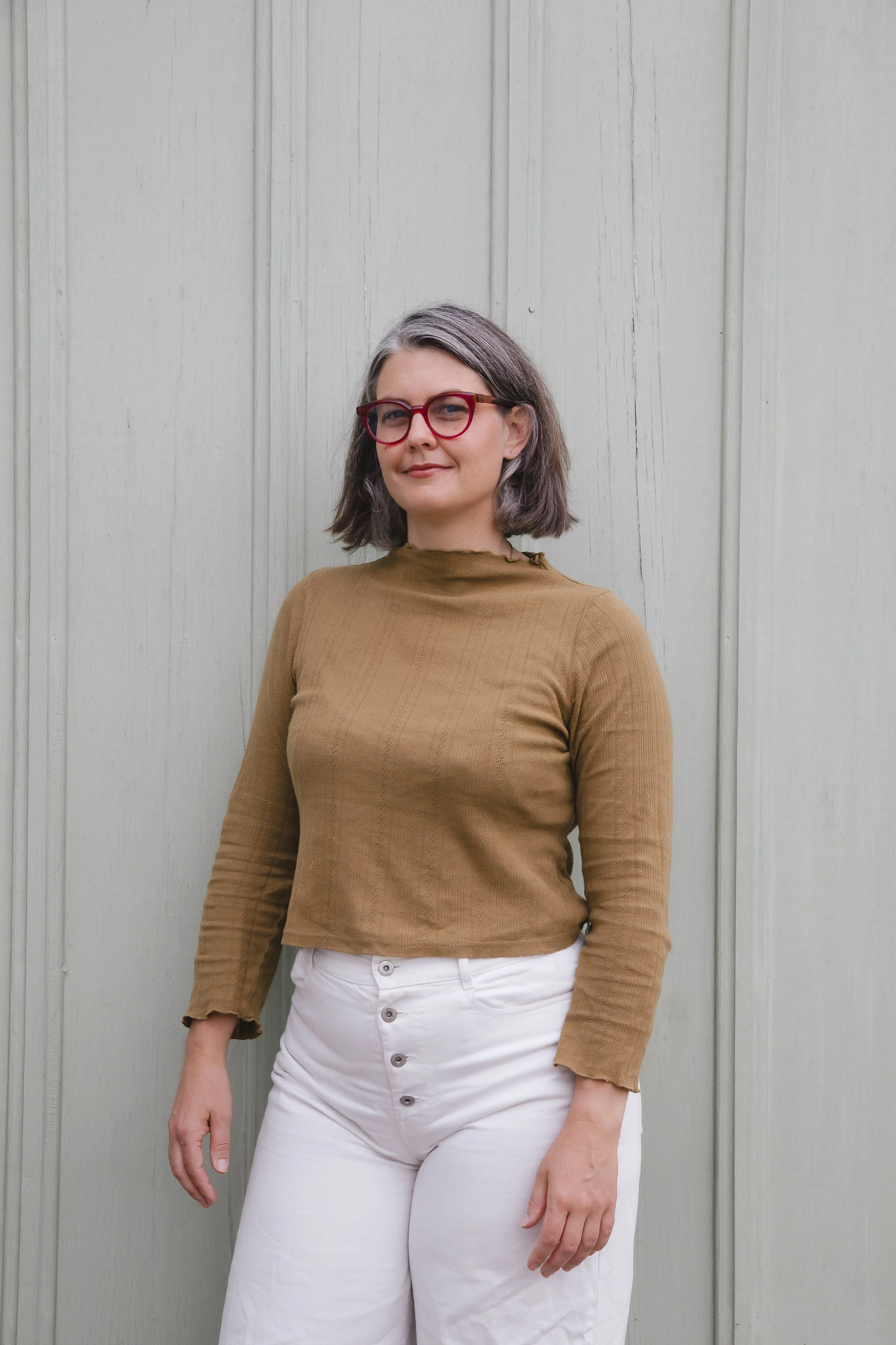 Willa stands in front of a white wall wearing red glasses, a dark yellow long sleeve shirt, and white jeans