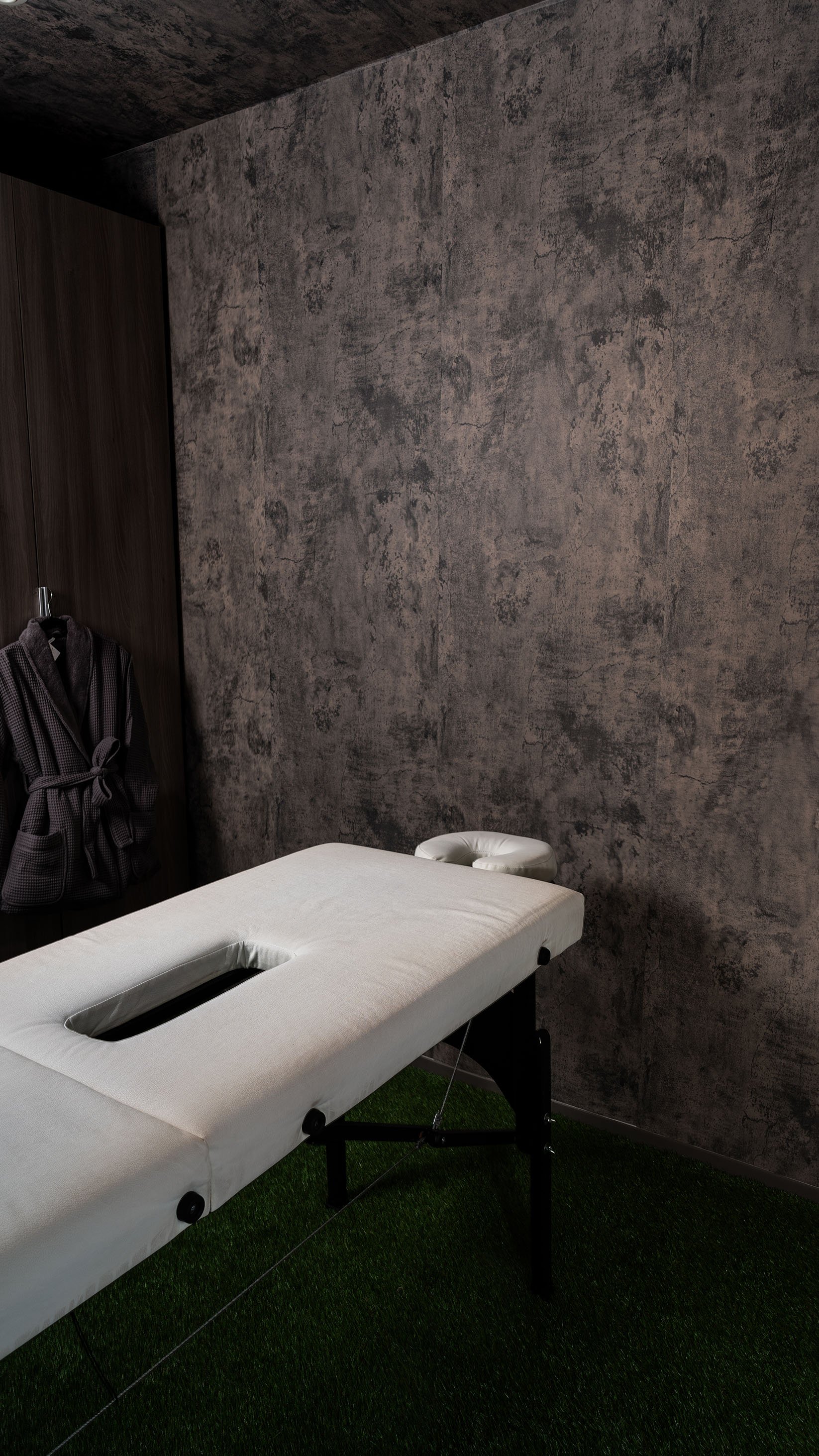 Cabina de masajes de estilo minimalista en Monterrey con camilla blanca, pared gris texturizada y piso de pasto sintético.