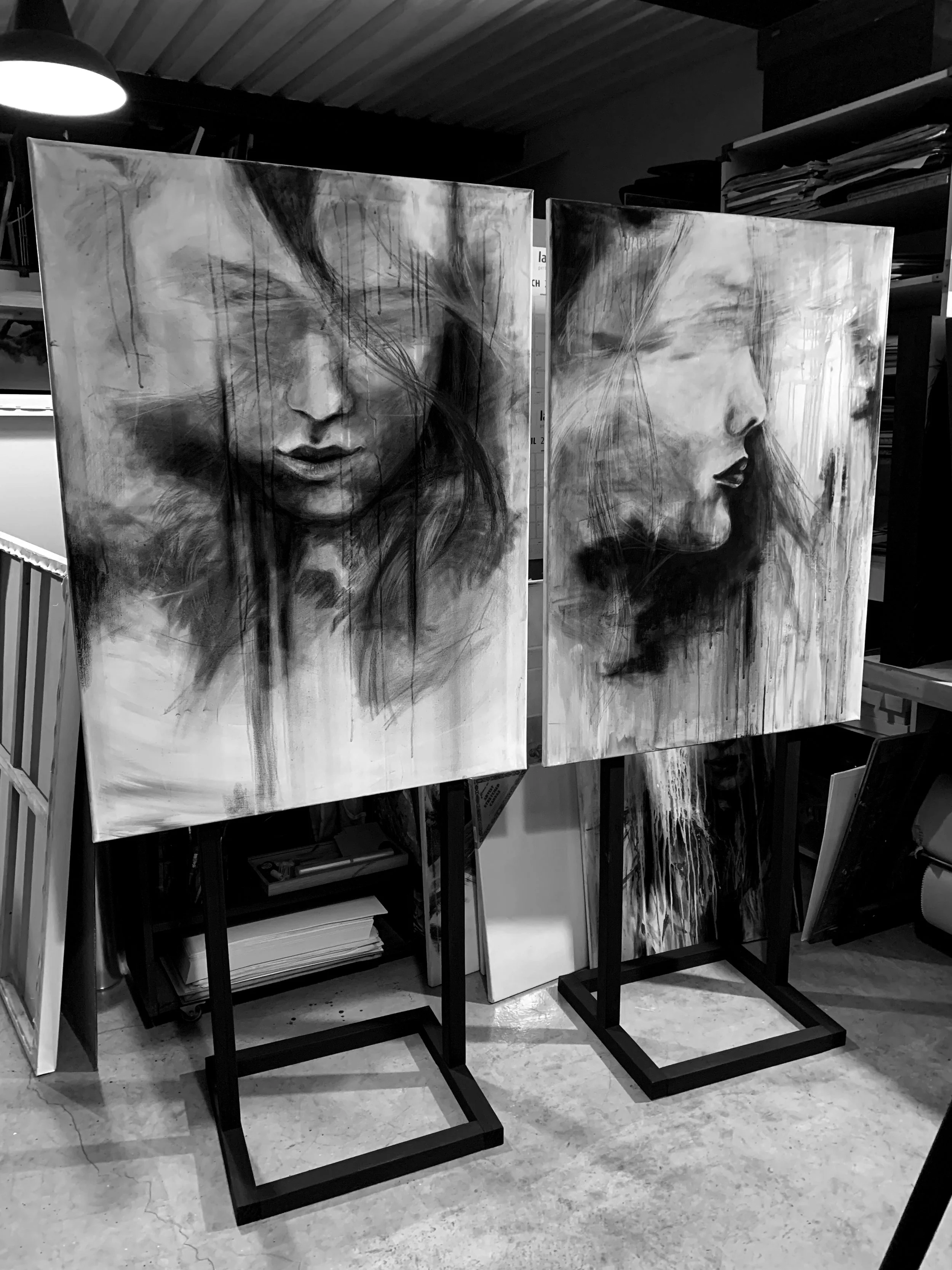 Two grayscale abstract portrait paintings of women on display, one showing a front view of a woman's face with eyes closed, hair flowing, and expression serene; the other showing a profile view with eyes open and lips slightly parted. Both paintings