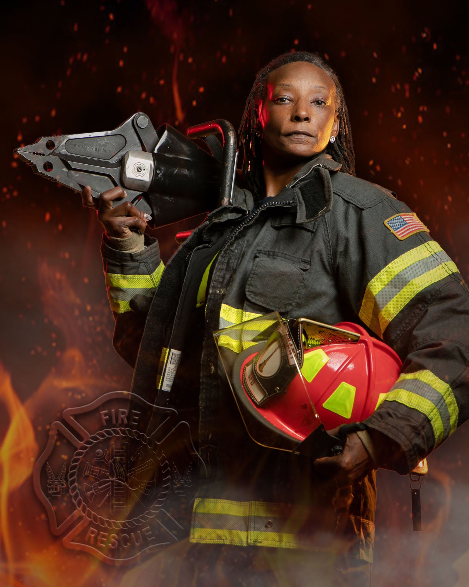 Firefighter in bunker gear holding her red helmet and a hydraulic rescue tool, flames in the background