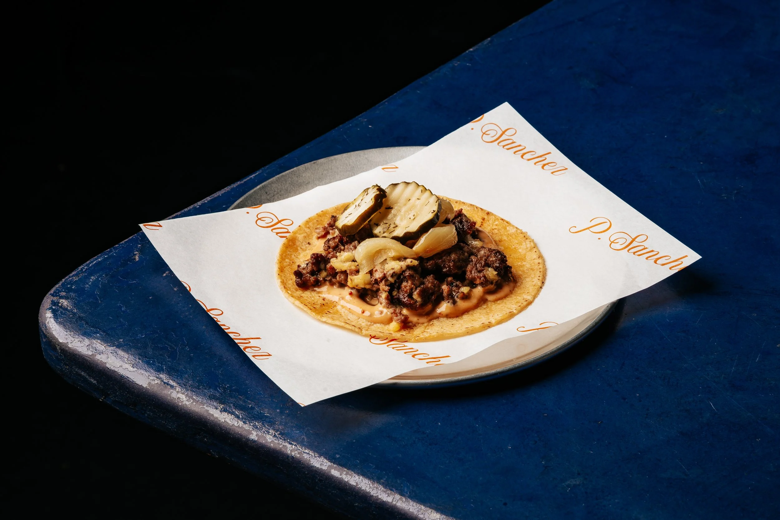 Taco with ground beef, cheese, pickles, and onions on a plate lined with branded paper from P. Sanch.