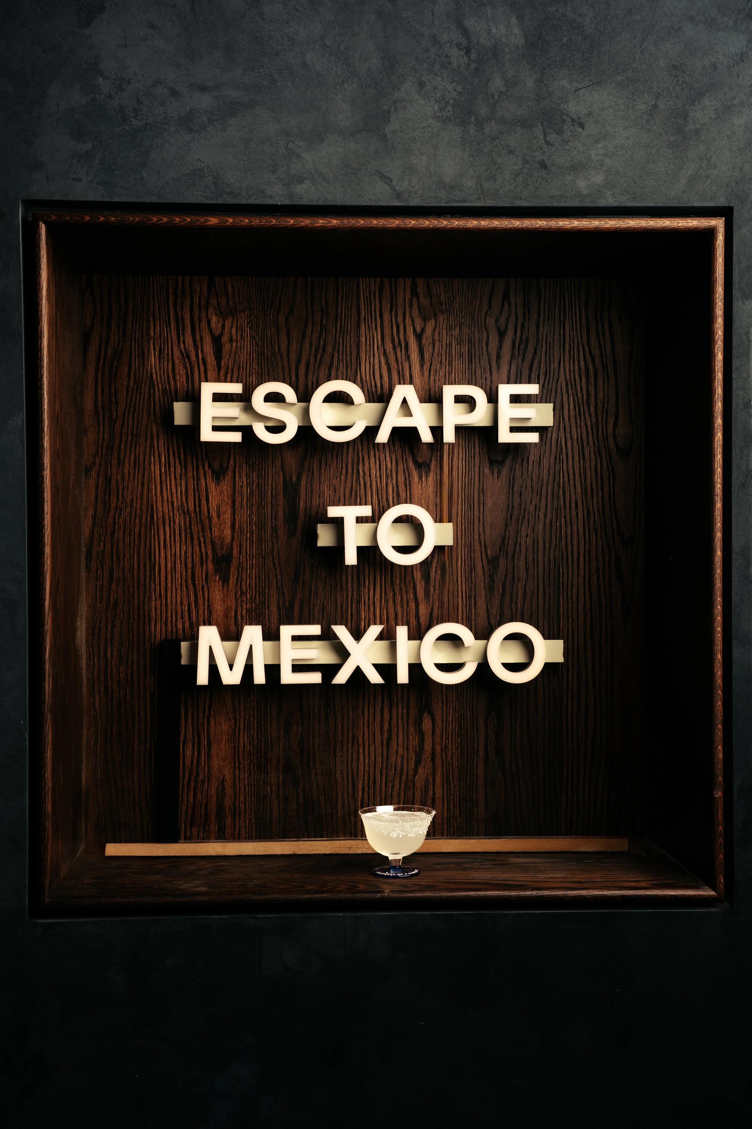 A wooden wall display with illuminated letters spelling 'ESCAPE TO MEXICO' and a margarita cocktail on the lower shelf.