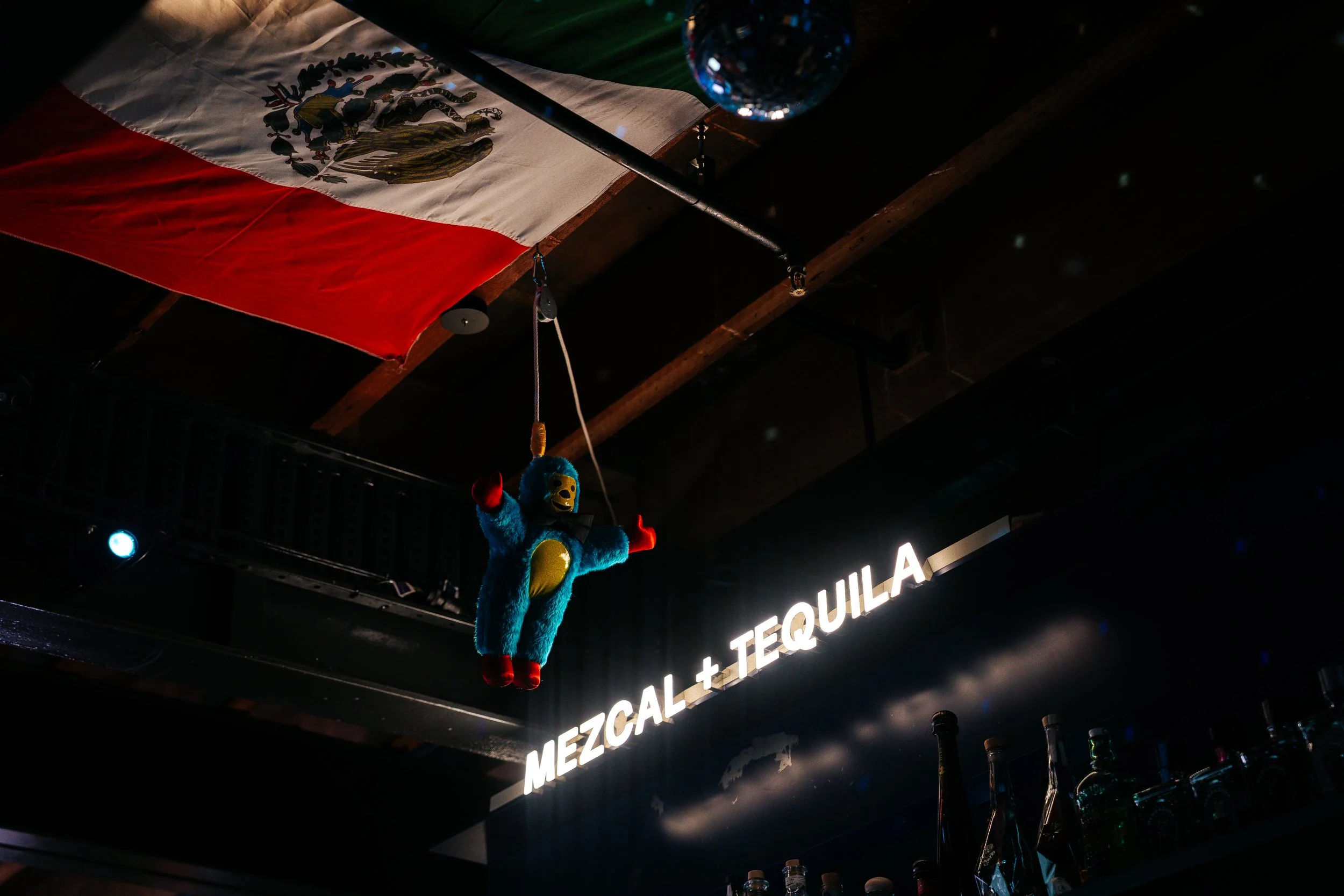 A Mexican flag hanging from the ceiling of a bar, with a plush toy of a blue character with a yellow face and red arms and legs hanging below it. Part of a neon sign reads 'MEZCAL + TEQUILA,' and bottles of alcohol are visible on a shelf below.