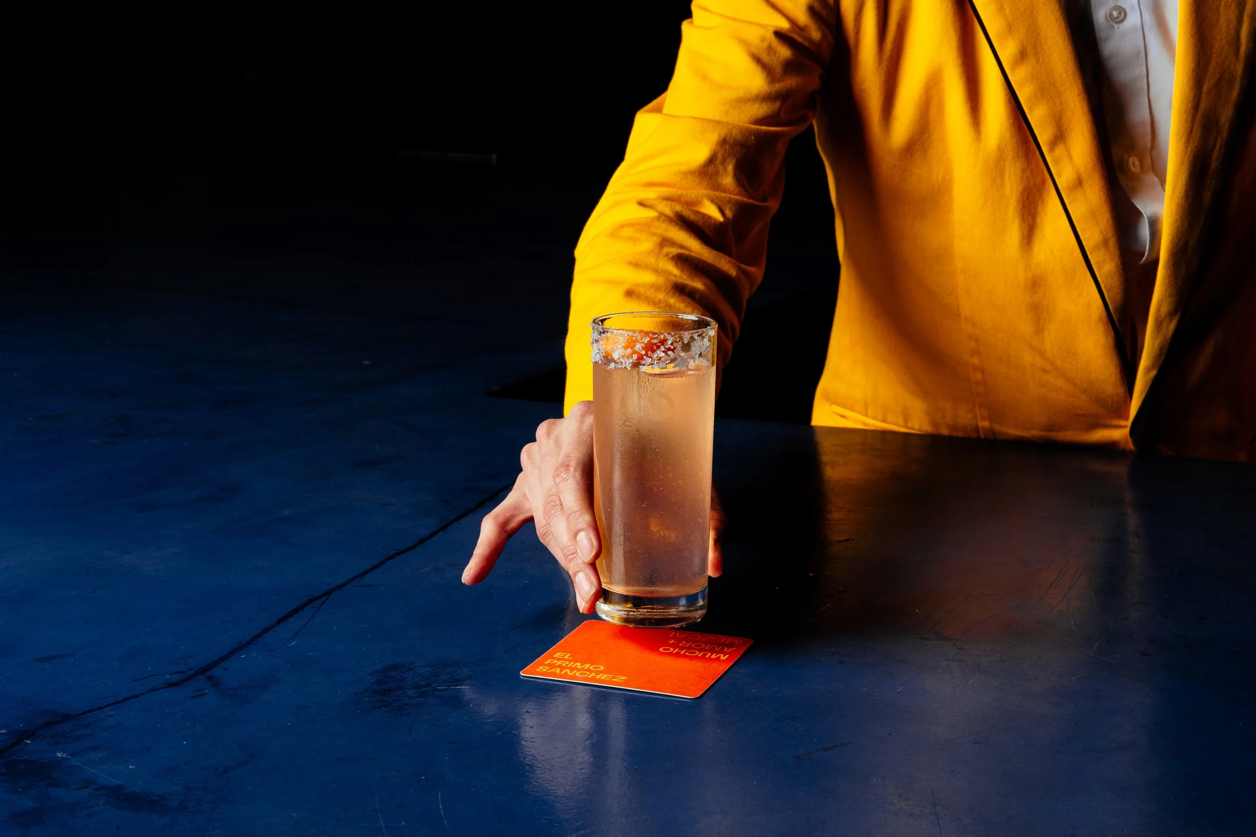 Person in a yellow jacket placing a glass of clear beverage on a red card labeled 'El Primo Sanchez' on a dark table.