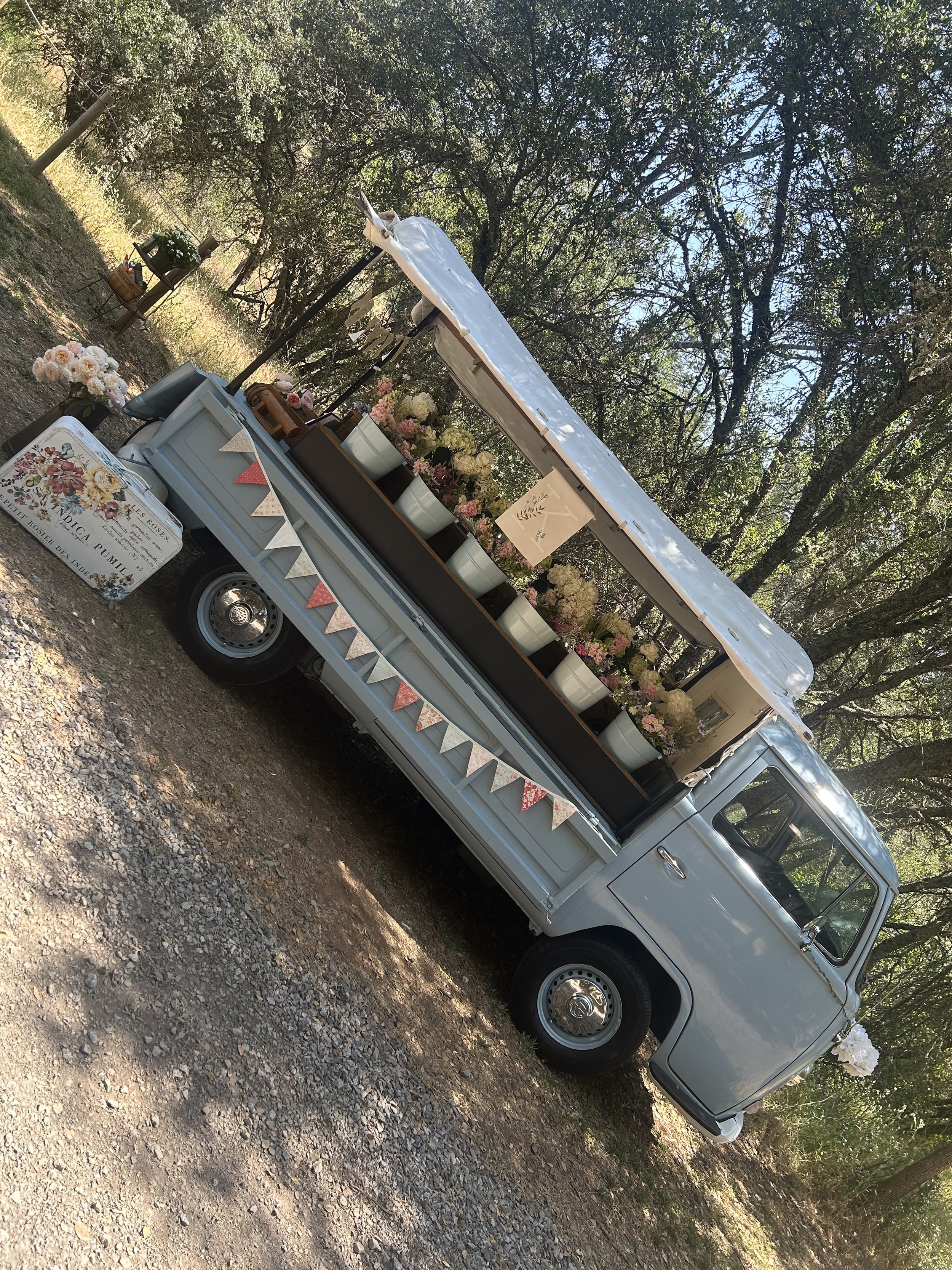 A vintage blue Volkswagon truck decorated with flowers and bunting, set outdoors with trees in the background.