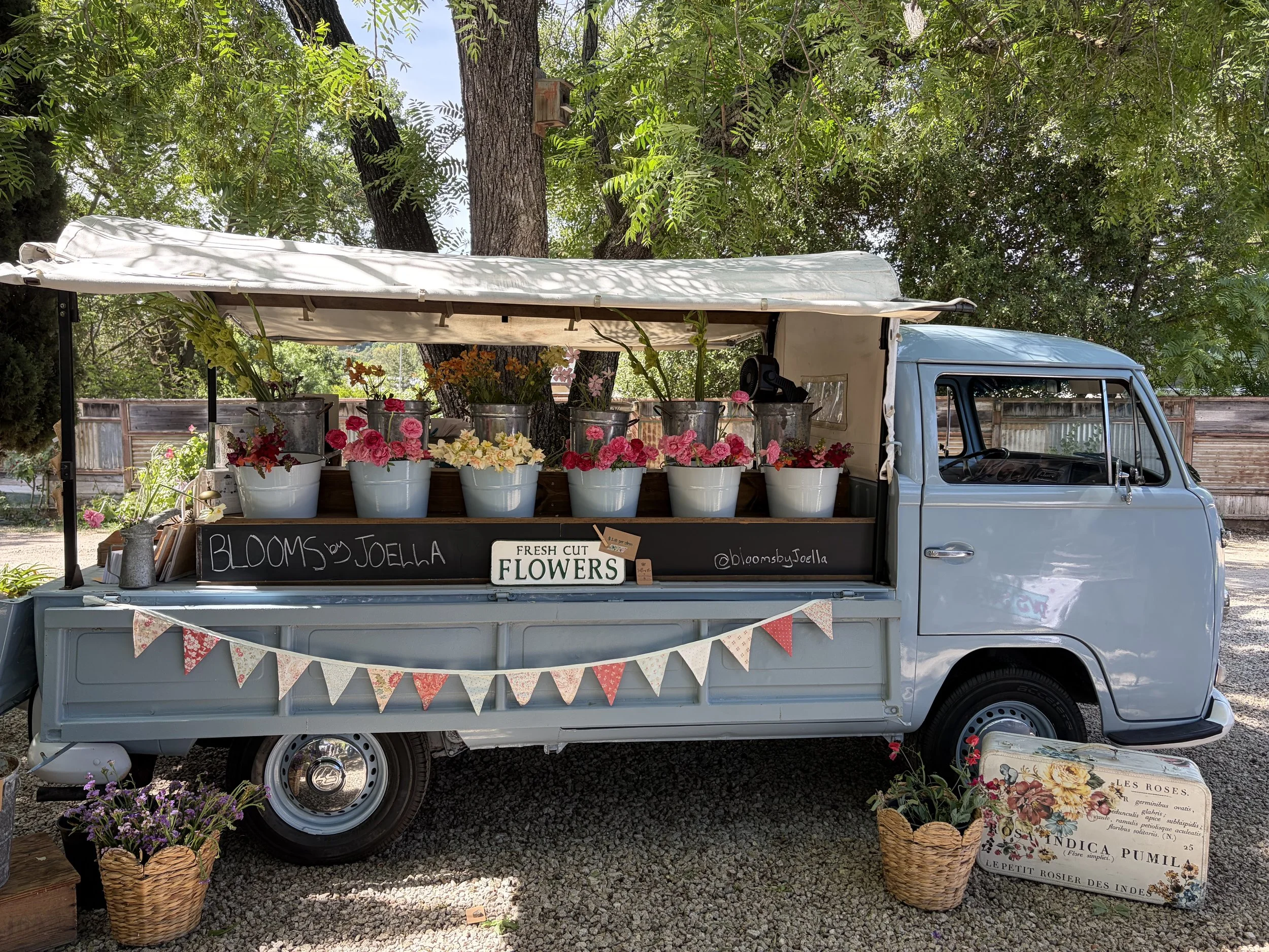 A vintage blue truck converted into a flower stand with buckets of pink and white flowers. The truck is parked outdoors under trees with a wooden fence in the background. There is a sign that reads 'BLOOMS by Joella' and another that says 'FRESH CUT FLOWERS.' Two wicker baskets with flowers sit on the ground next to the truck.