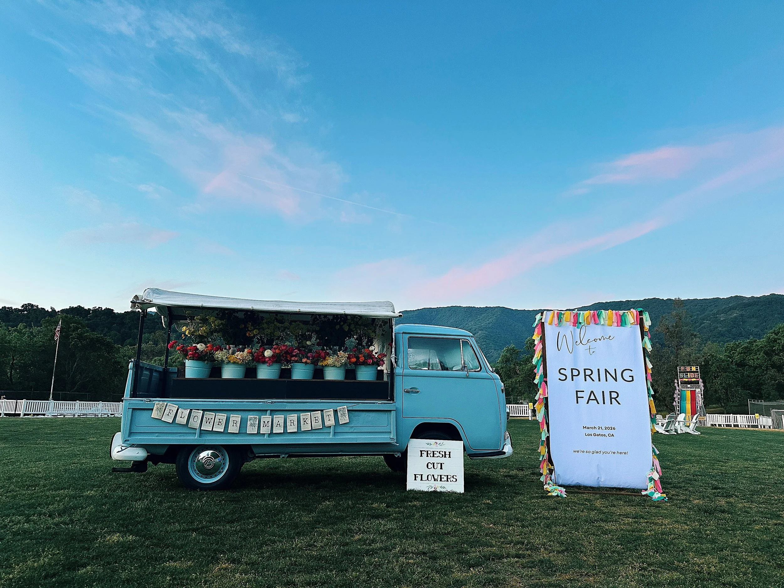 A vintage blue truck decorated with flowers and a sign reading 'Flower Market' is parked on a grassy field during daytime. Next to it, a large welcoming sign announces a spring fair on March 21, 2026, in Los Gatos, California. The scene includes a white fence in the background and colorful decorations around the signs.