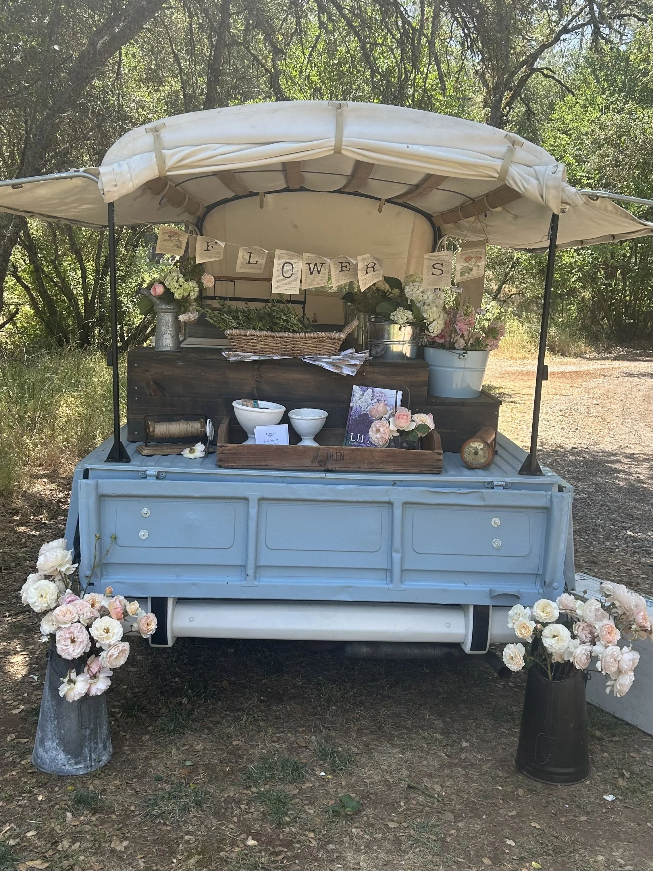 A small blue vintage pickup truck with a canopy, decorated with flowers and a banner that spells 'FLOWERS,' set outdoors among trees and greenery.