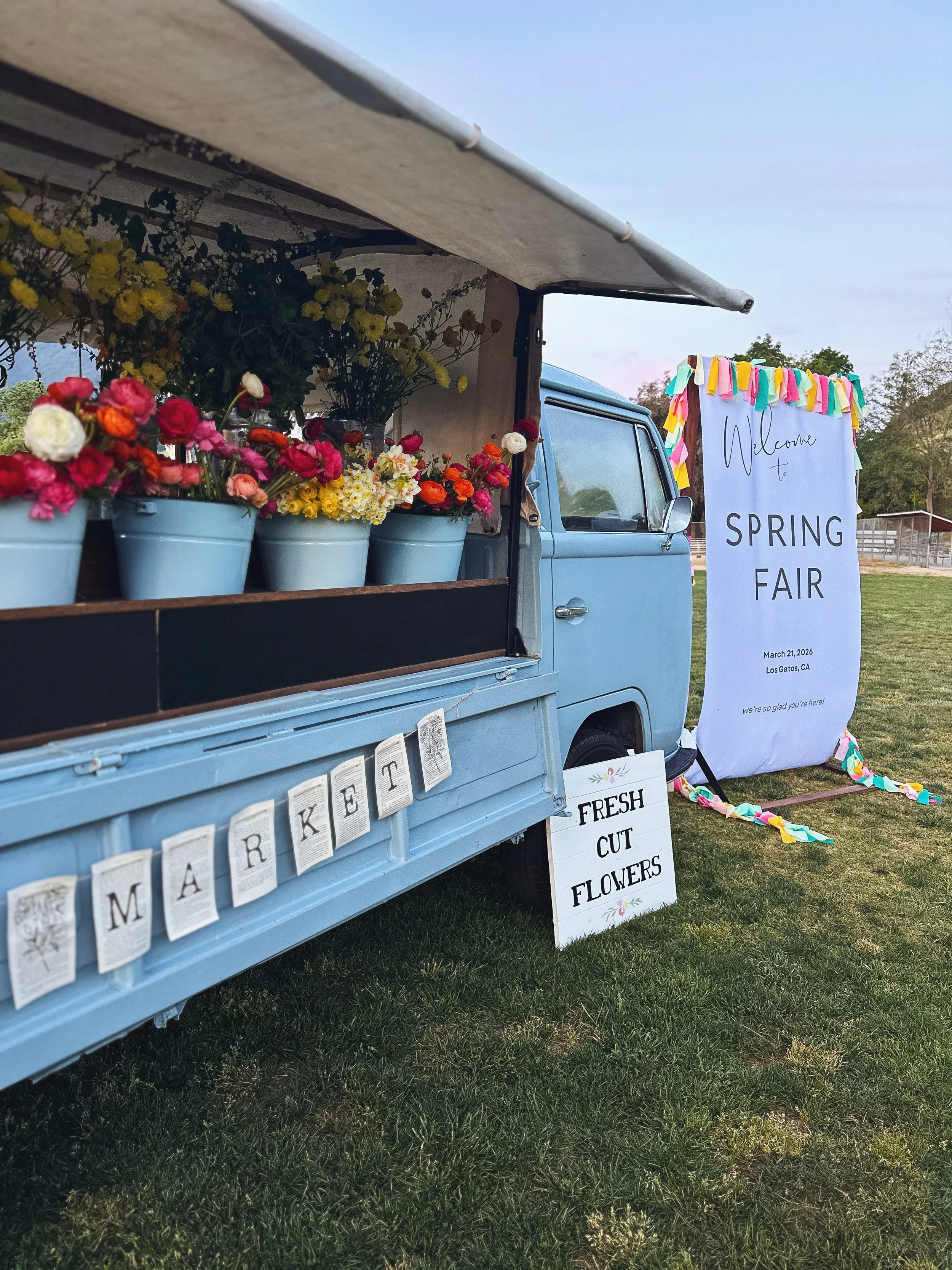 A vintage blue truck with a flower stand selling fresh cut flowers, and a white sign that reads 'Welcome to Spring Fair,' March 21, 2026, Los Gatos, CA.