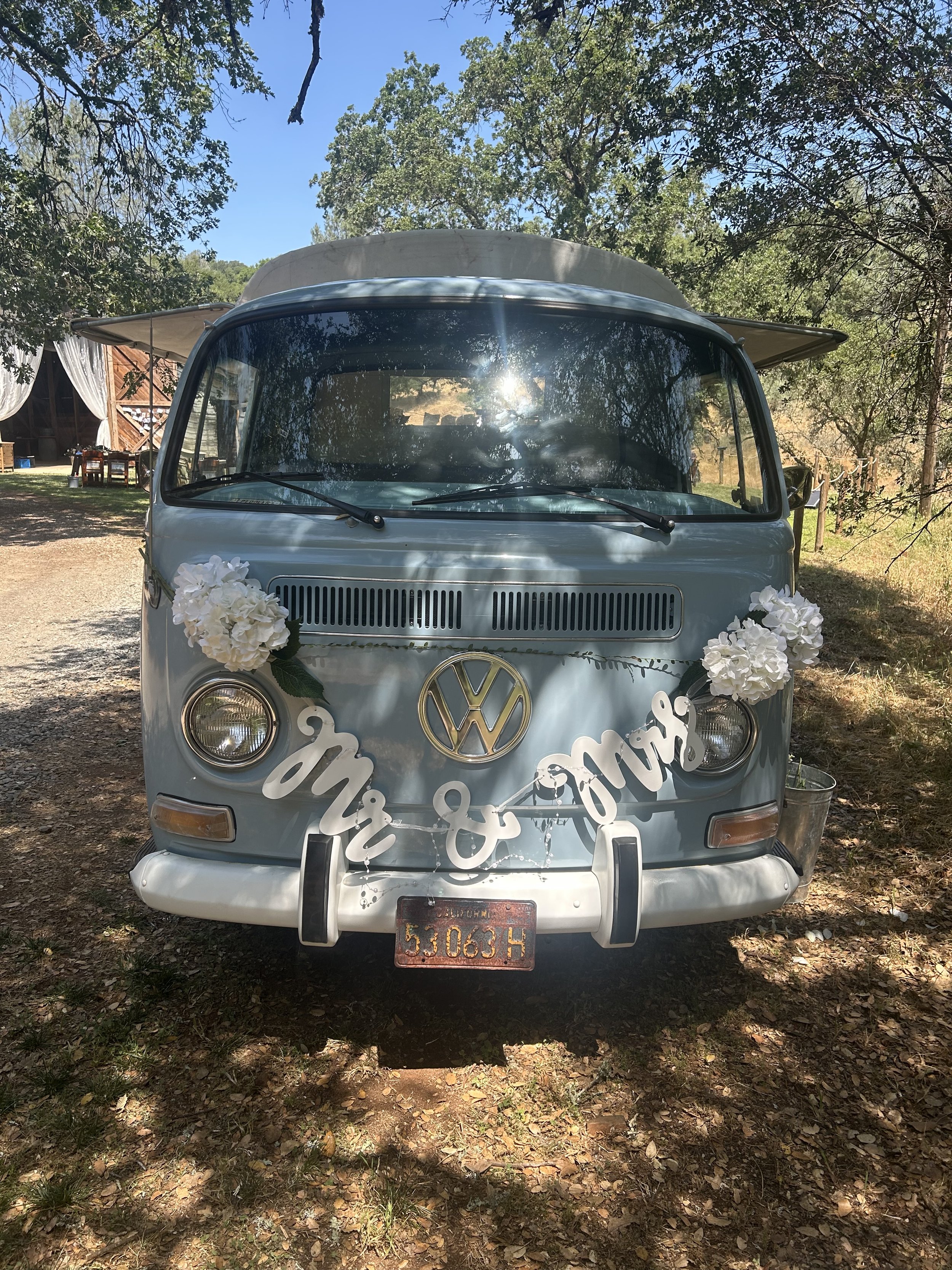 A vintage Volkswagen truck decorated with white flowers and a 'Mr & Mrs' sign, parked outdoors under trees during daytime.