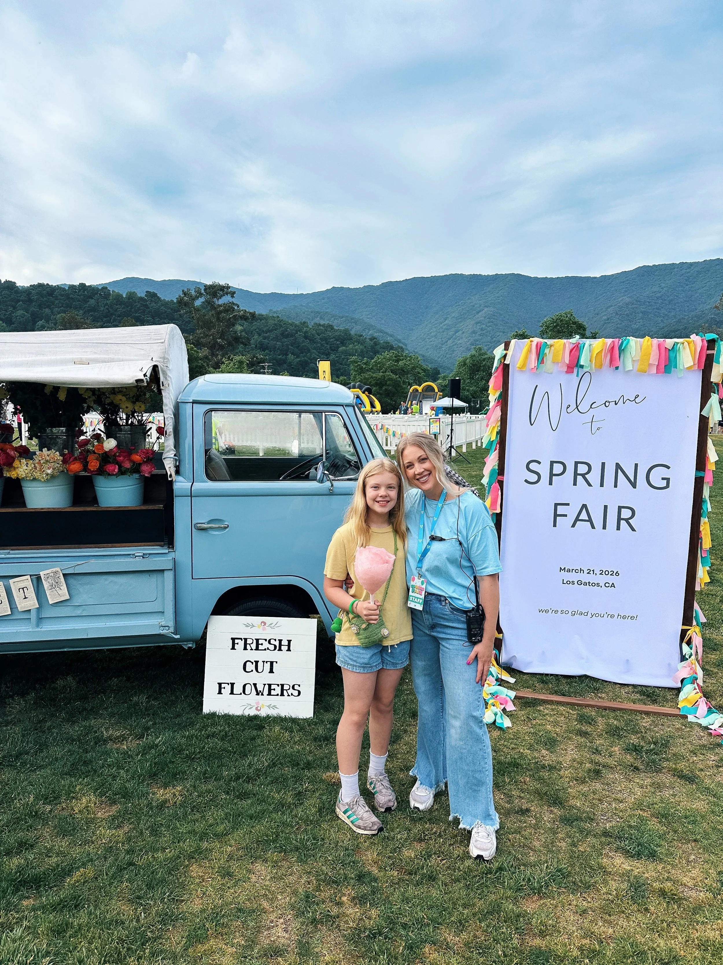 A woman and a girl smiling at a spring fair, standing in front of a sign that reads 'Welcome to Spring Fair' with mountains in the background. The girl holds cotton candy, and there is a vintage blue truck with flowers and a sign that says 'Fresh Cut Flowers' nearby.