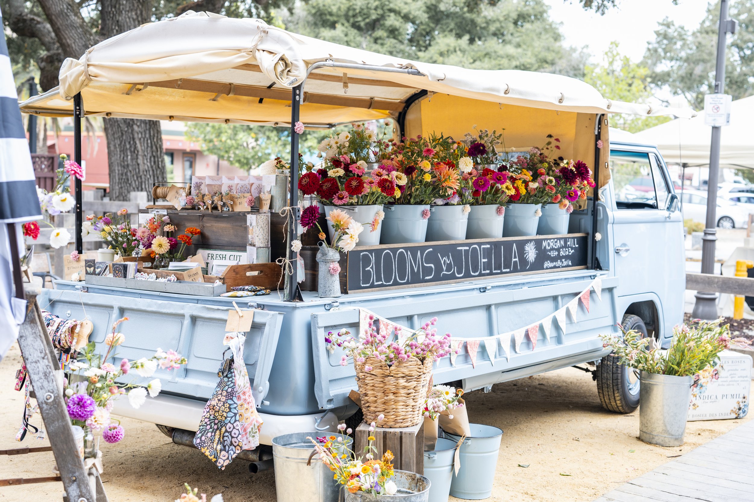A vintage blue truck with a yellow canopy selling colorful flowers and small plants at an outdoor market.