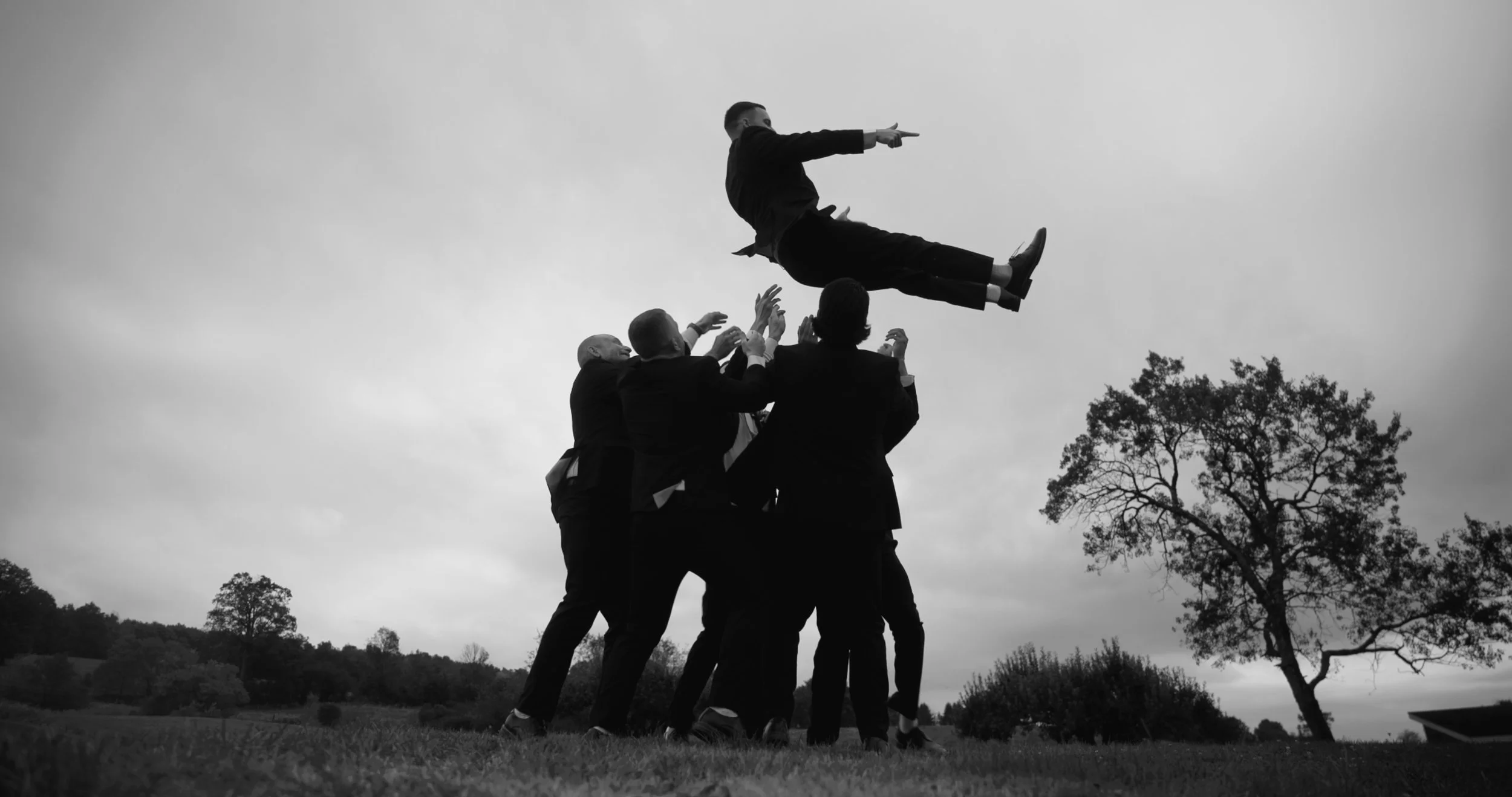 A group of men in suits are holding up a man in a suit who is floating or flying in the air with arms extended outward, as if in a superhero pose, outdoors against a cloudy sky with trees in the background.