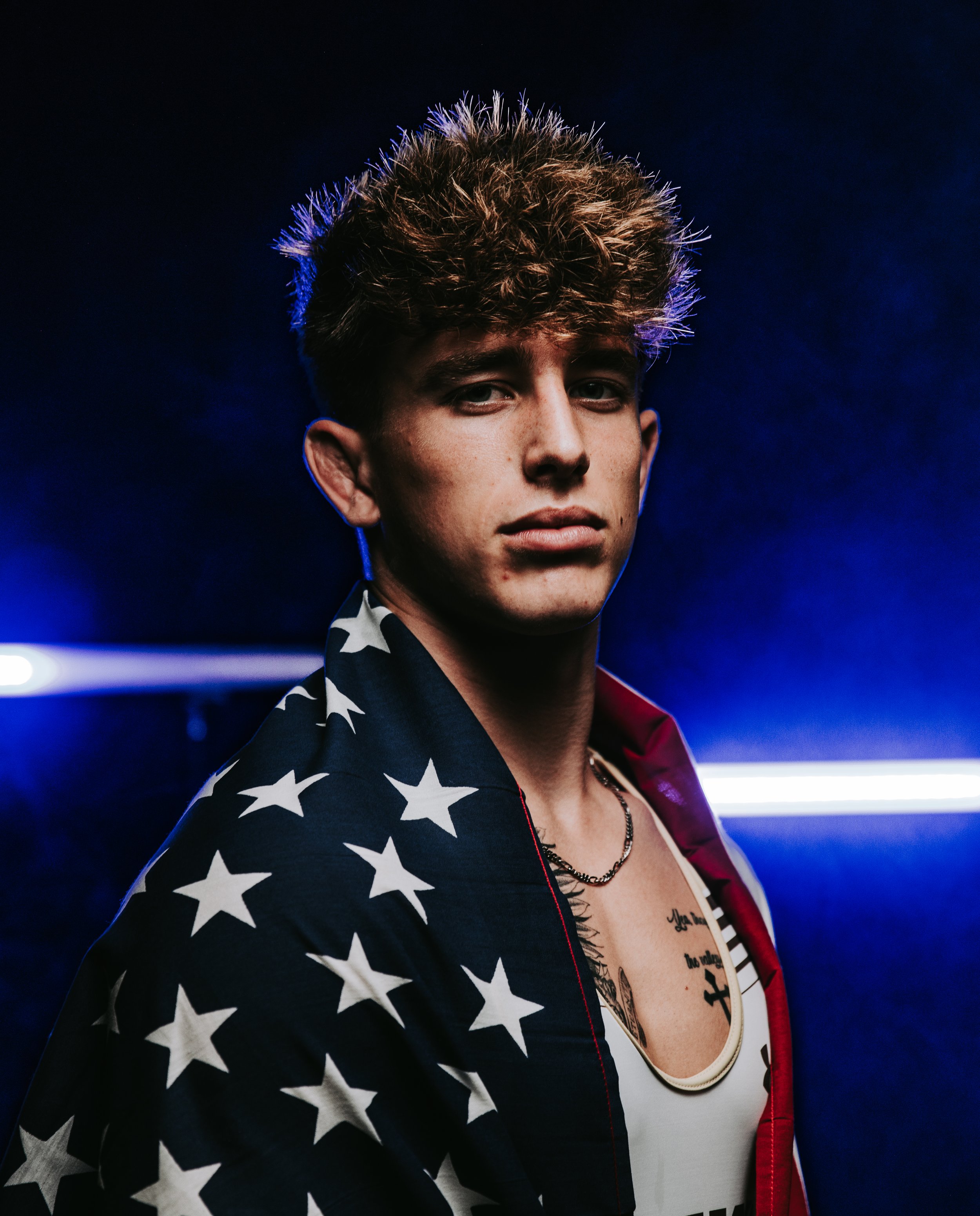 Close-up of a young man with tan skin and curly light brown hair, wearing a jacket with a stars pattern, a patchwork-style shirt, and a necklace, standing against a dark background with blue lights.
