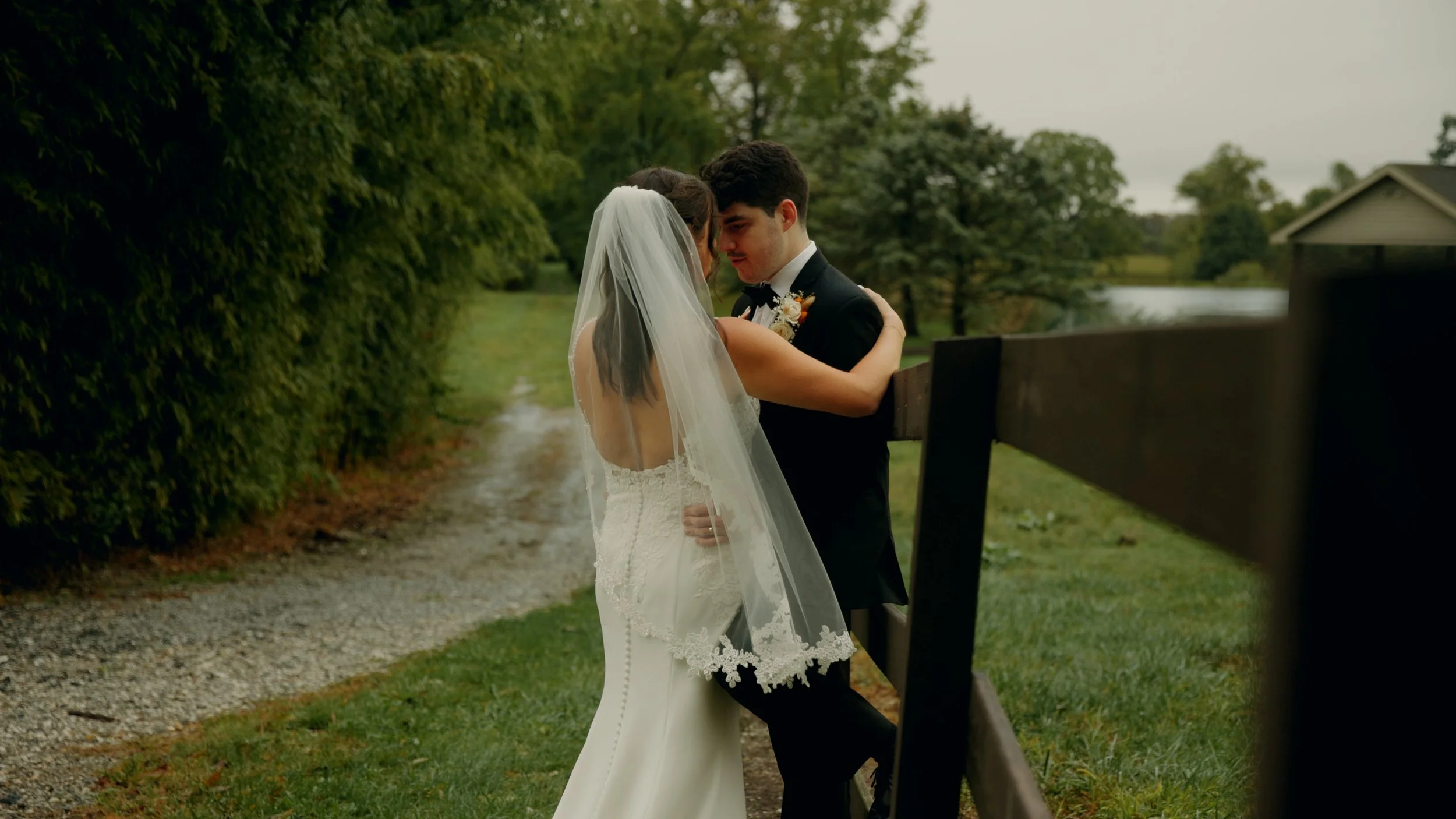 A bride and groom sharing a romantic moment outdoors, standing close with their foreheads touching, near a wooden fence, with trees and a cloudy sky in the background.