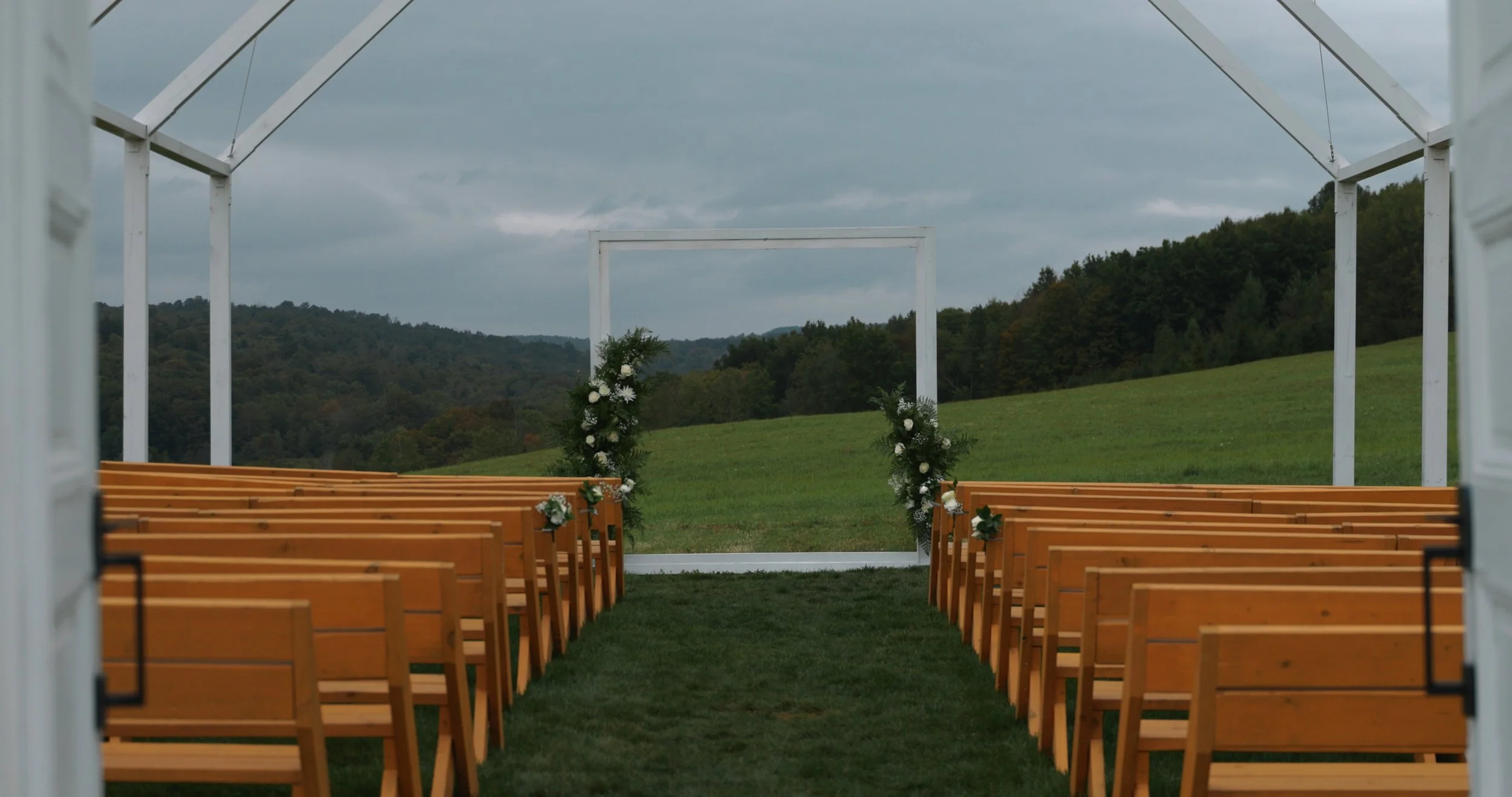 Outdoor wedding ceremony setup with wooden benches, decorated with flowers, facing a white arch with greenery and flowers on a grassy area, hills and cloudy sky in the background.