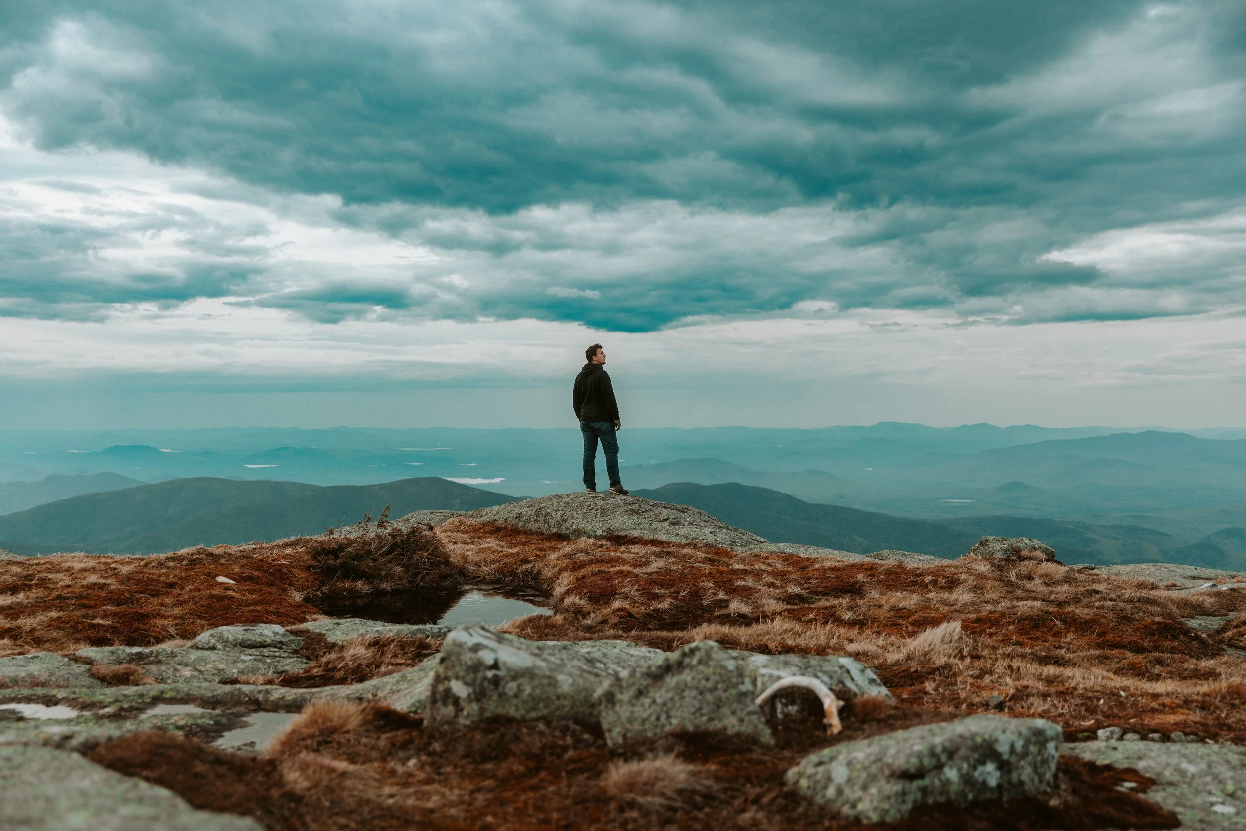 A person standing on a rocky mountain ledge, looking out at a landscape of rolling hills with a lake in the distance under a cloudy sky.