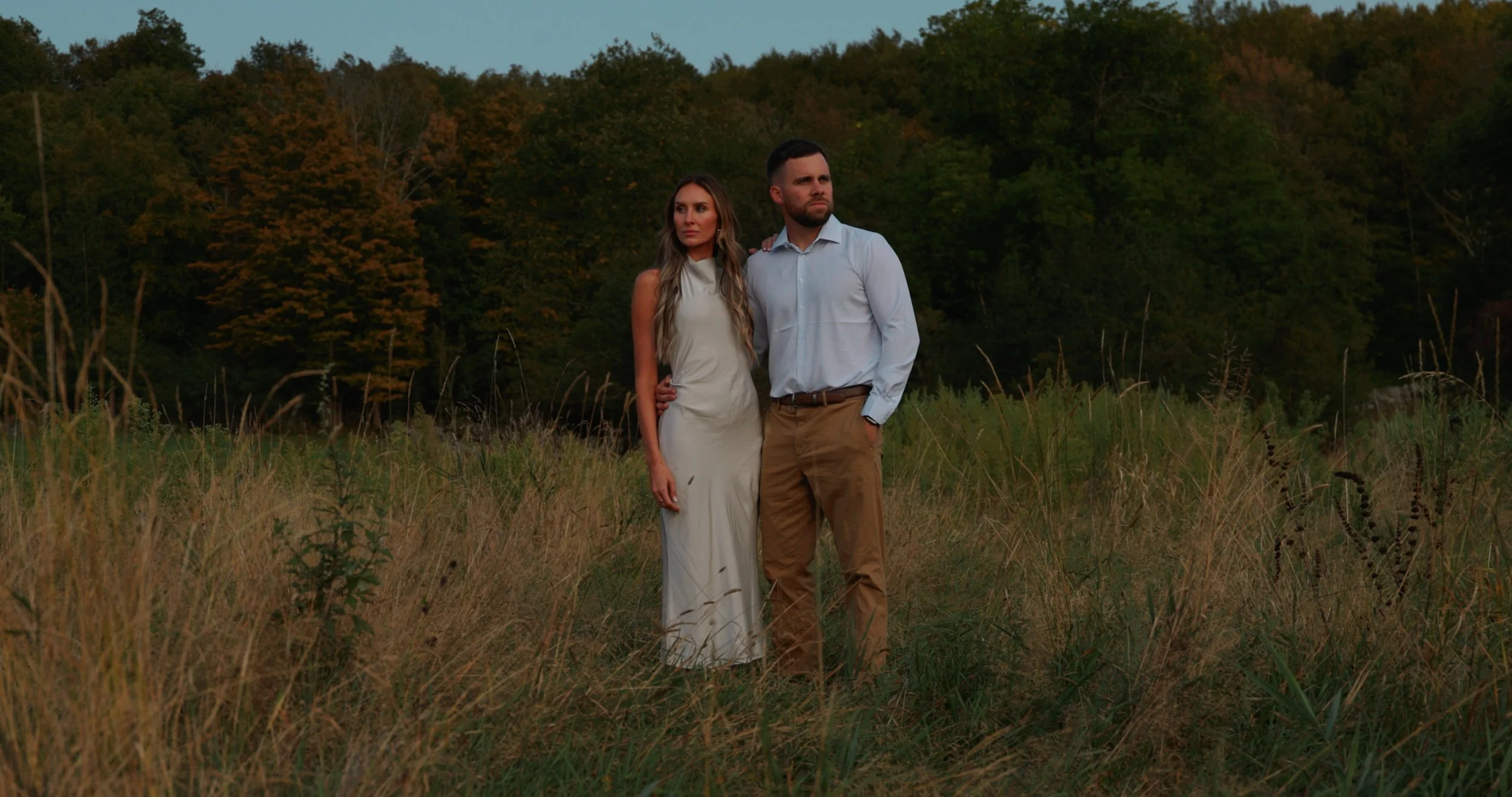A man and woman standing close together in a grassy field with trees in the background during twilight. The woman is wearing a white dress, and the man is wearing a light blue shirt and khaki pants.