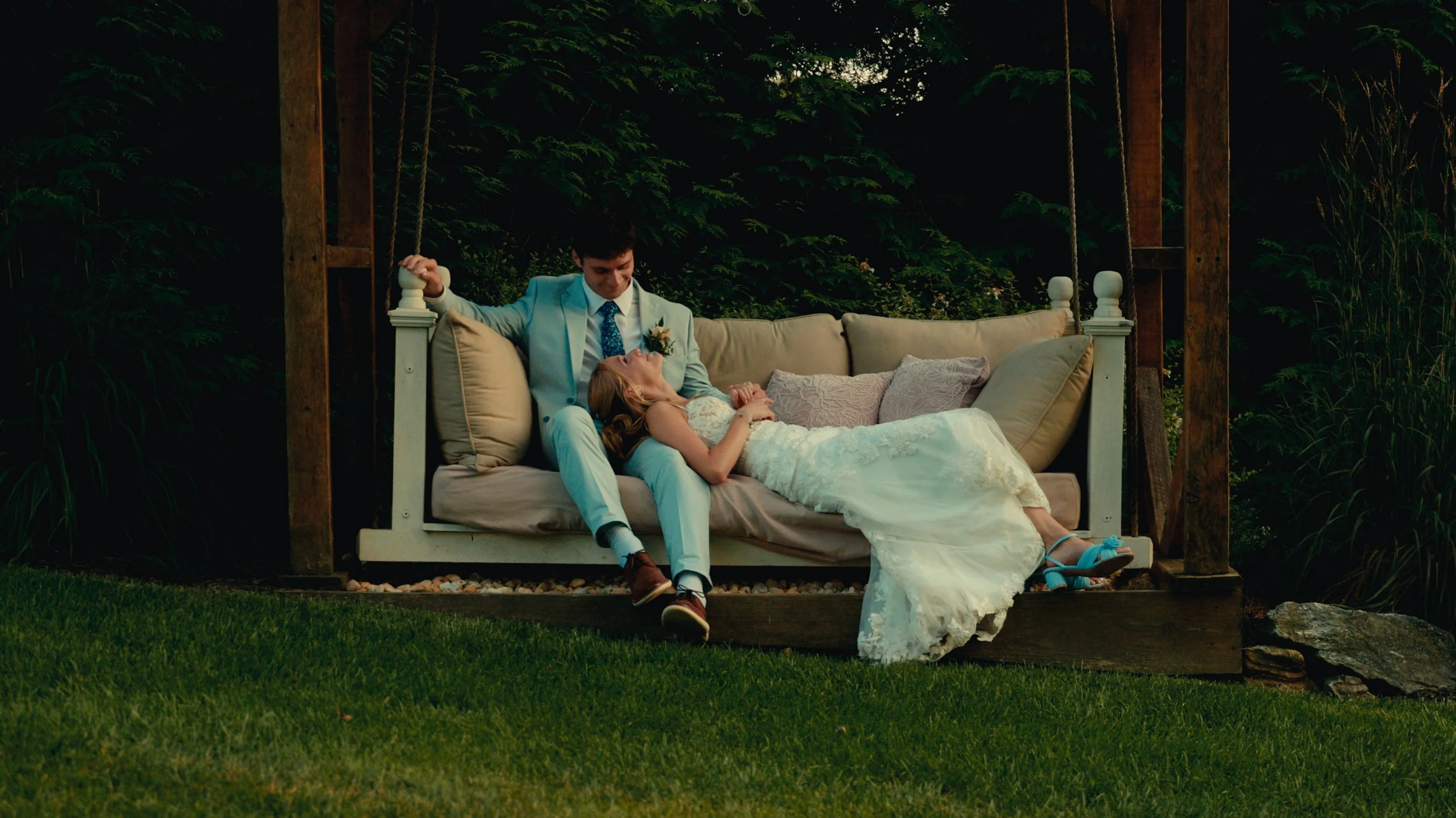 A groom in a light blue suit is sitting on a swing bed, leaning back with a woman in a white wedding dress lying on his lap, both smiling, in a lush garden setting.