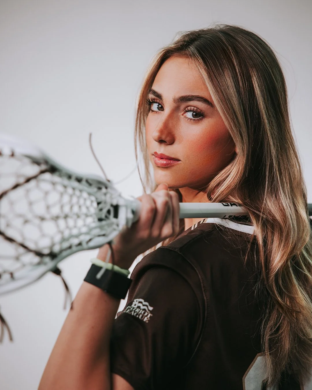 Close-up of a young woman with long, wavy hair holding a lacrosse stick over her shoulder, wearing a sports jersey, looking confidently at the camera.