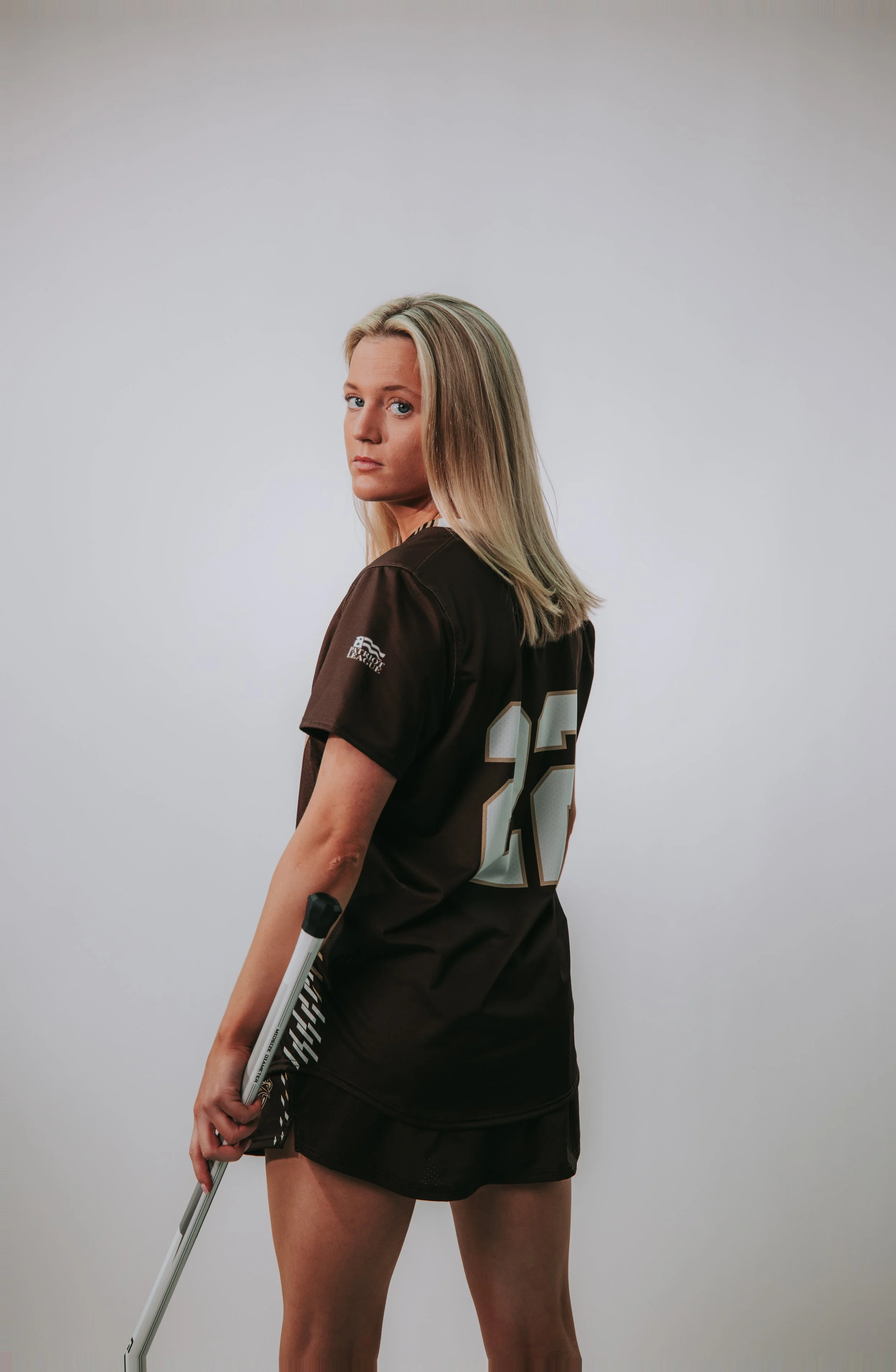 A woman with long blonde hair wearing a black sports jersey with the number 23 on the back, holding a hockey stick, looking over her shoulder at the camera, standing against a plain gray background.