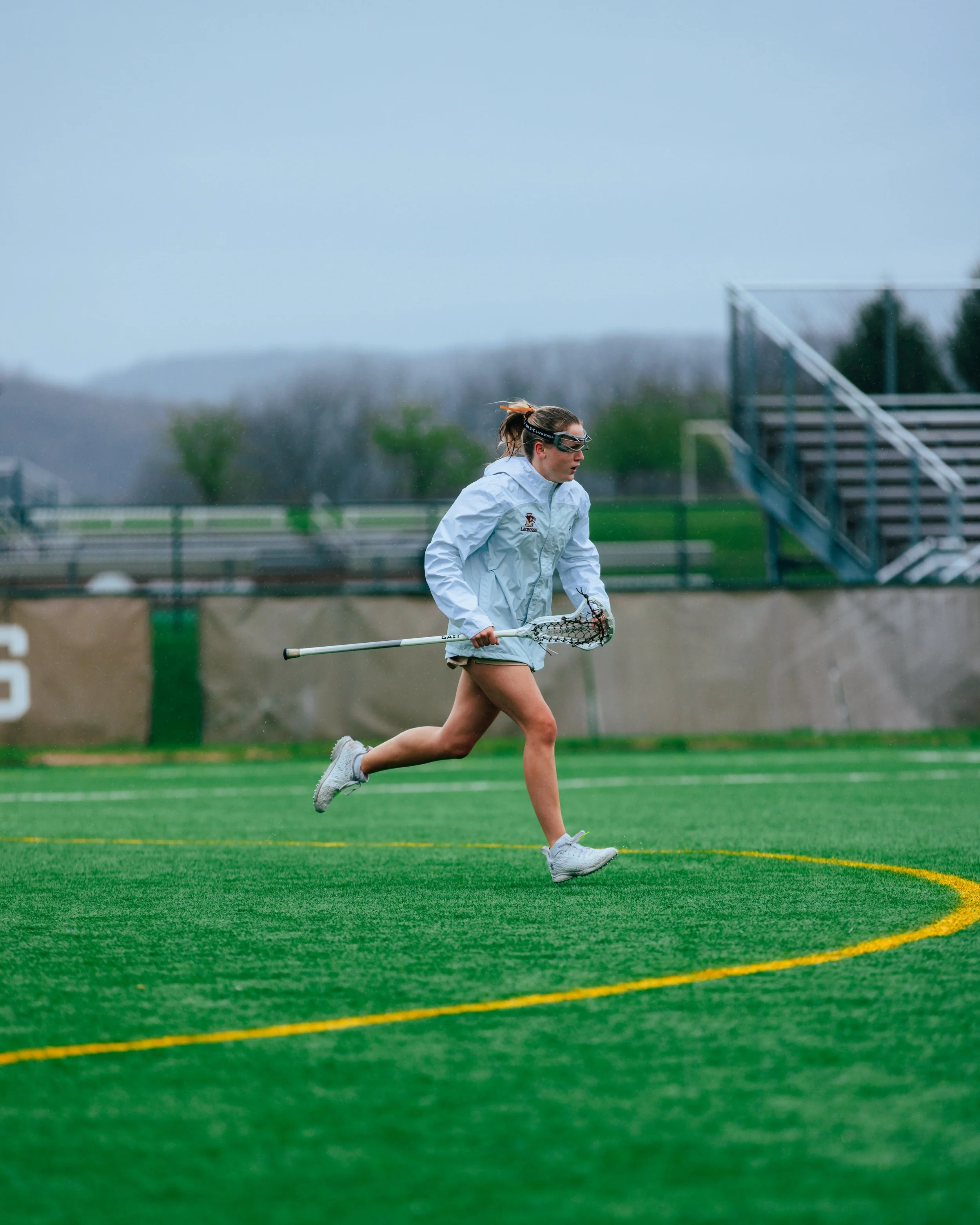 A female lacrosse player running on a green field, holding a lacrosse stick, with a cloudy sky and bleachers in the background.