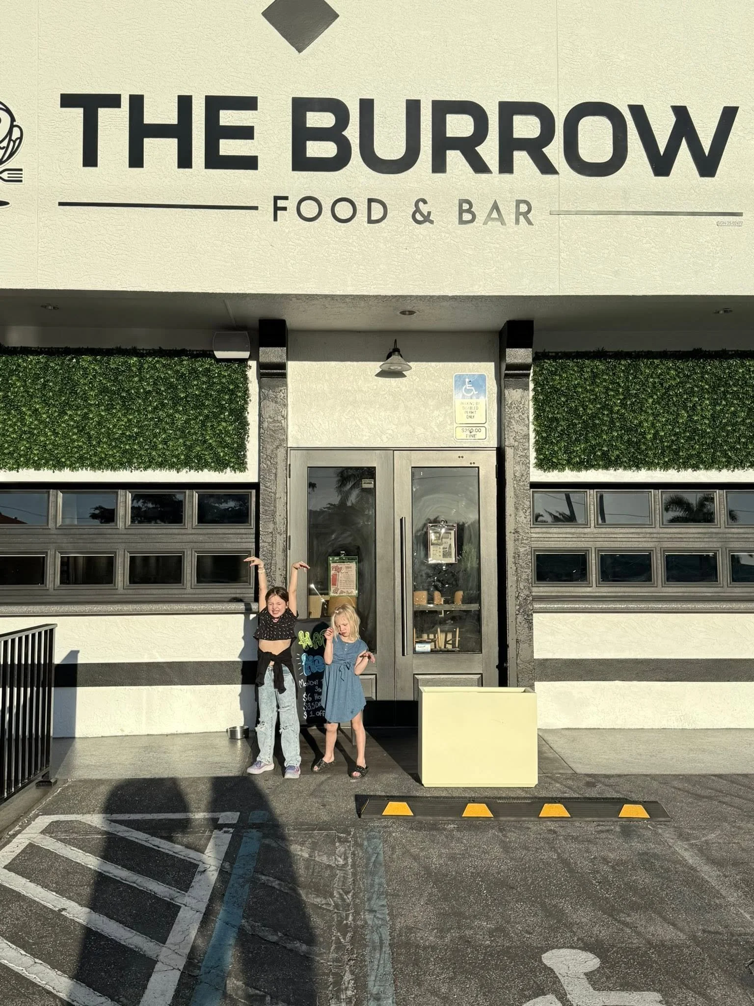 Two young girls standing outside the entrance of a restaurant called The Burrow Food & Bar, with one girl raising her arms and smiling.