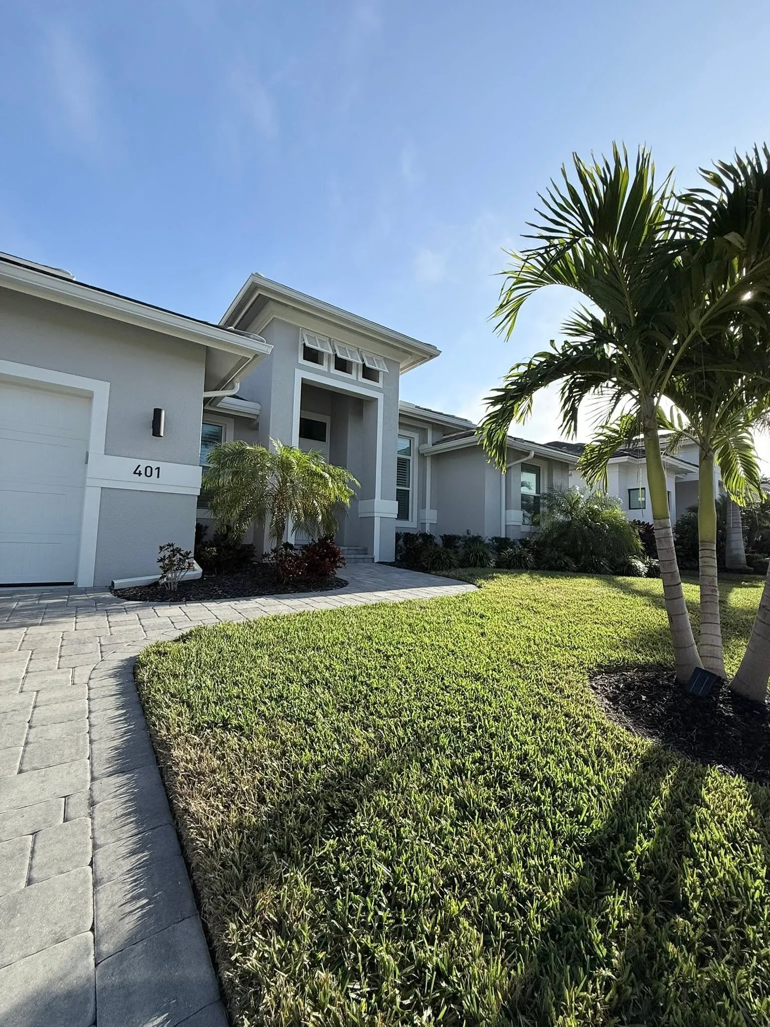 Front yard of a modern house with a neatly manicured lawn, a paved walkway, and tropical palm trees under a clear blue sky.