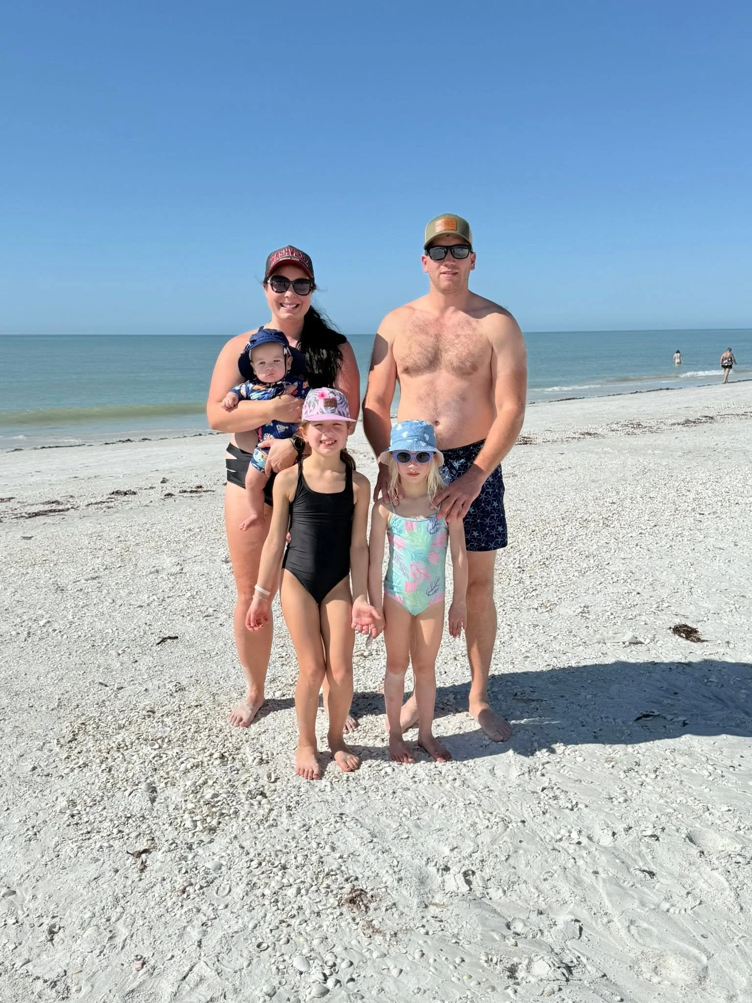 A family of five on a sandy beach with calm ocean waves and a clear blue sky. Two adults and three children stand together, smiling at the camera.