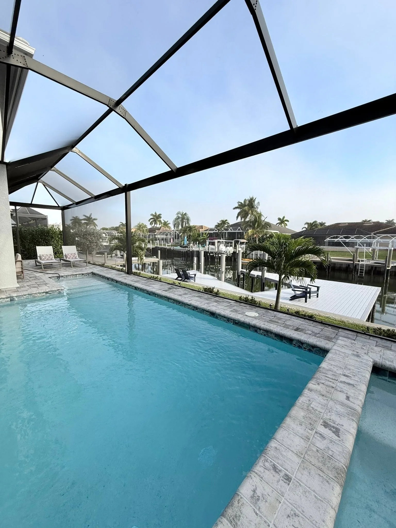 A backyard swimming pool with a screened enclosure, surrounded by lounge chairs, palm trees, and a canal with houses and boat docks in the background.