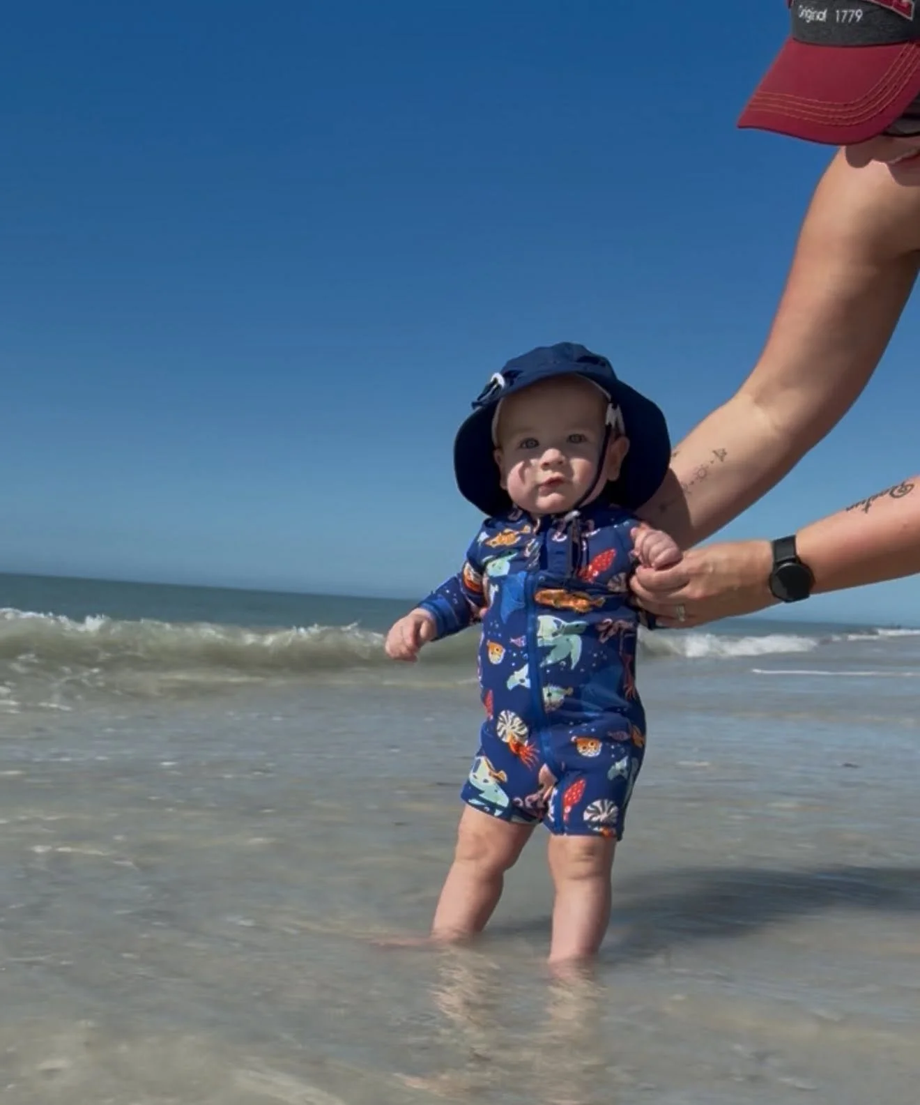 A young child in a colorful, ocean-themed swimsuit and sun hat standing in shallow water at the beach, held by an adult whose arm is extended from the right, with waves in the background under a clear blue sky.